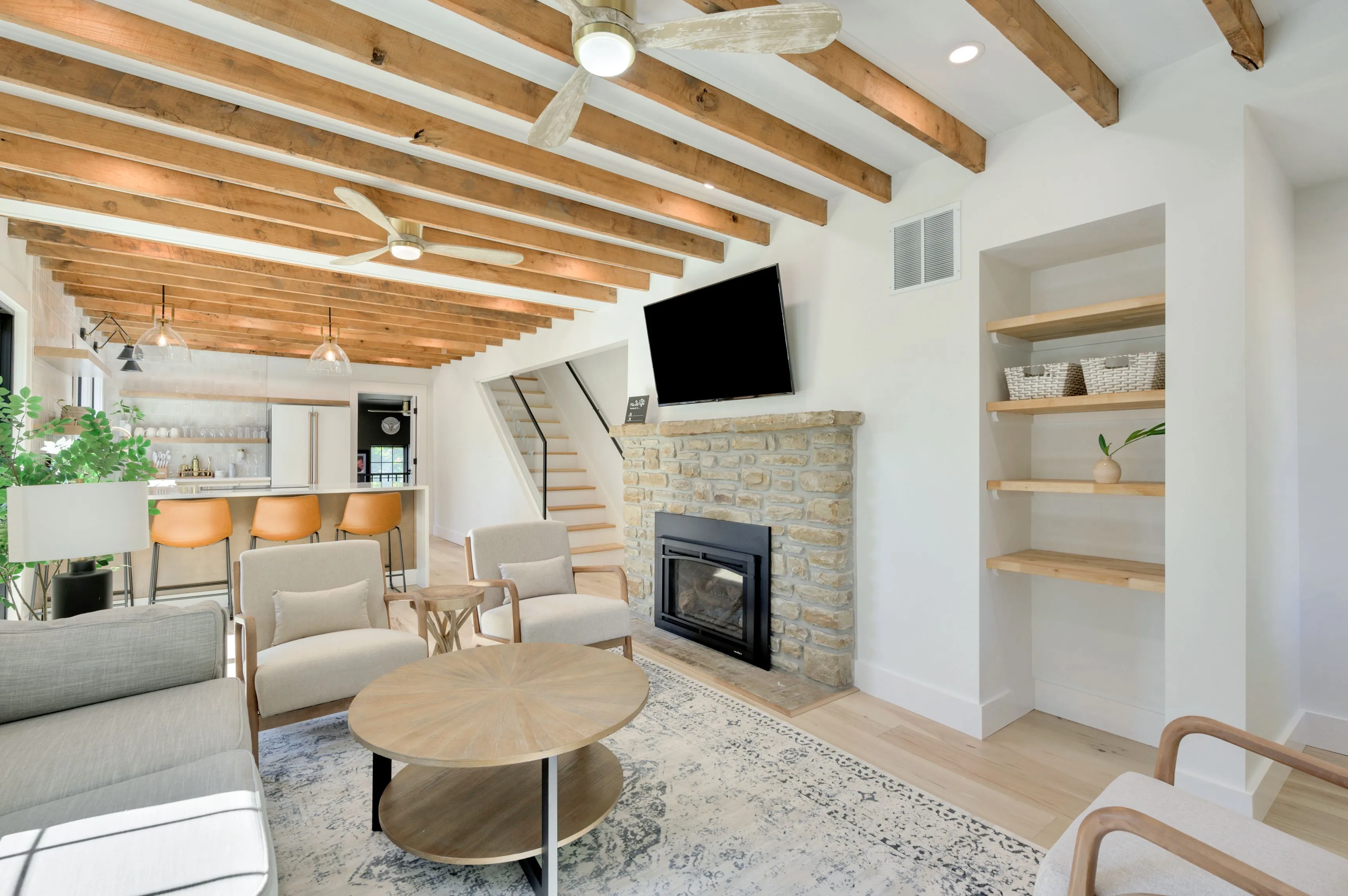 Modern living room with exposed wooden beams, a fireplace, built-in shelving, and a staircase leading to a loft.