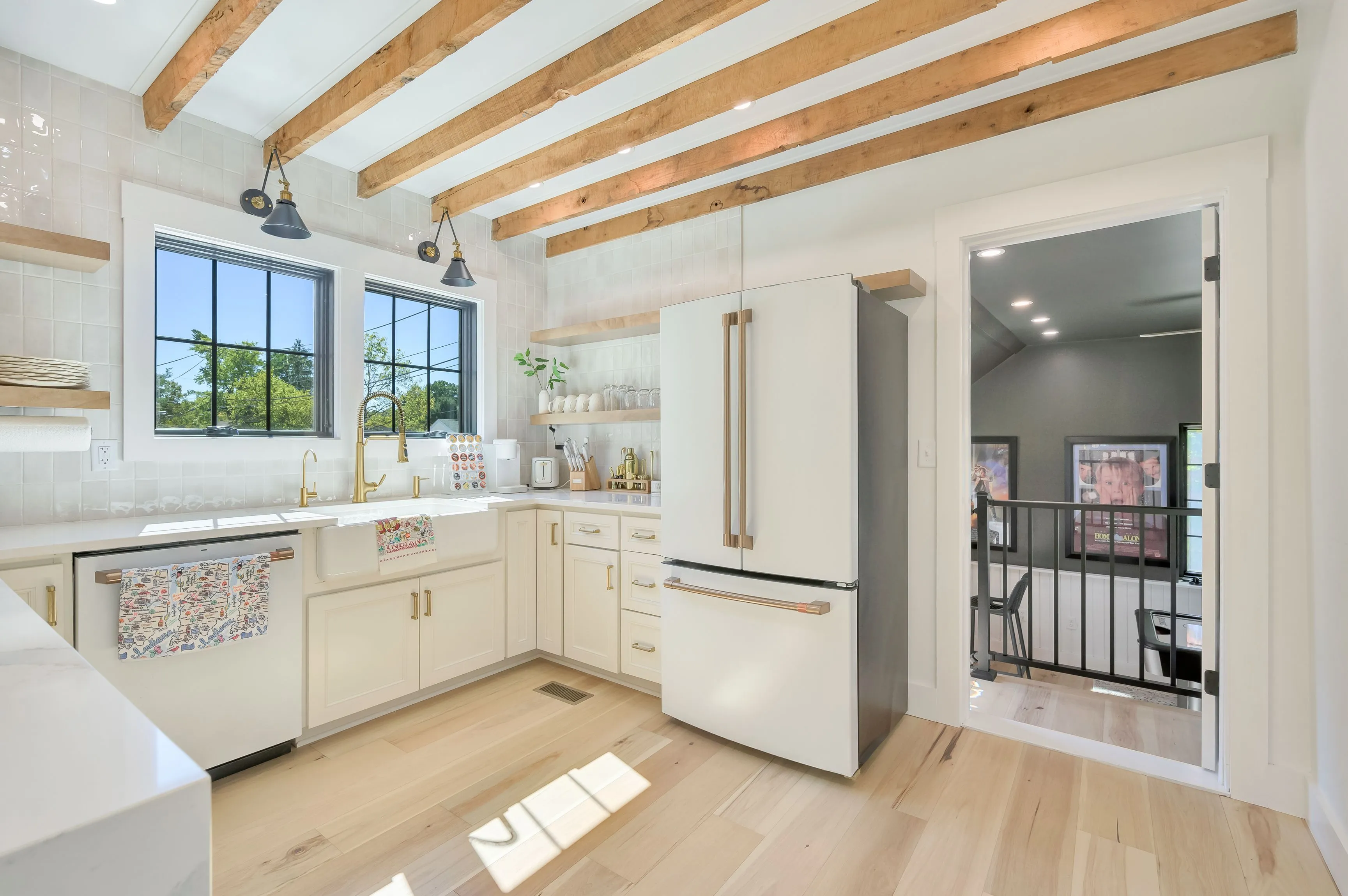 Bright modern kitchen with wooden beams, white cabinetry, stainless steel appliances, and an open doorway leading to an adjacent room.