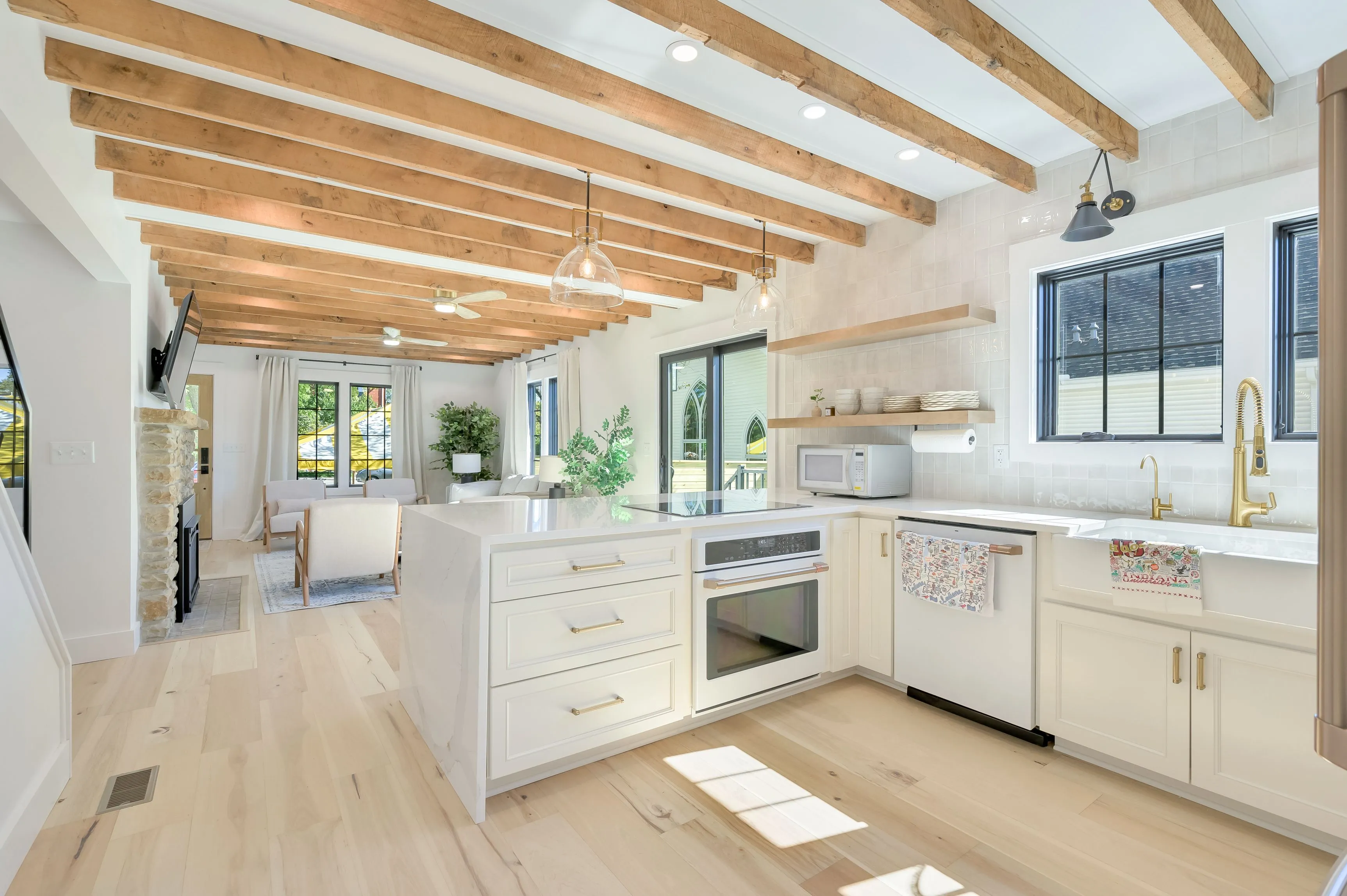 Bright and airy modern kitchen with white cabinetry, marble countertops, and exposed wooden beams leading into an open-plan living area with large windows.