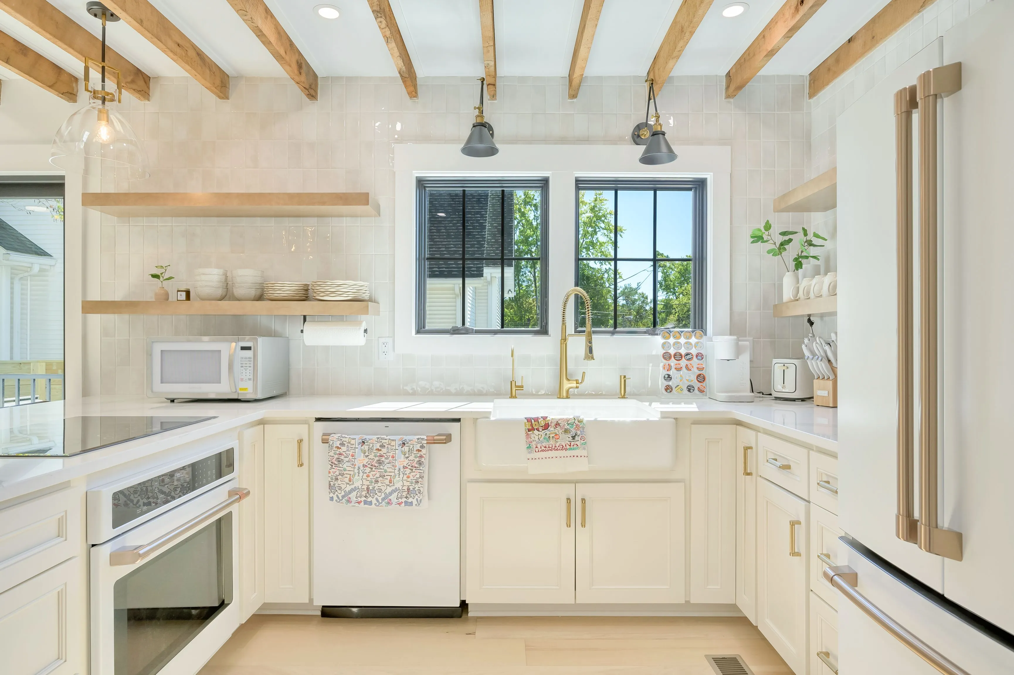 Bright and airy kitchen interior with white cabinetry, a farmhouse sink, floating wooden shelves, and exposed ceiling beams.