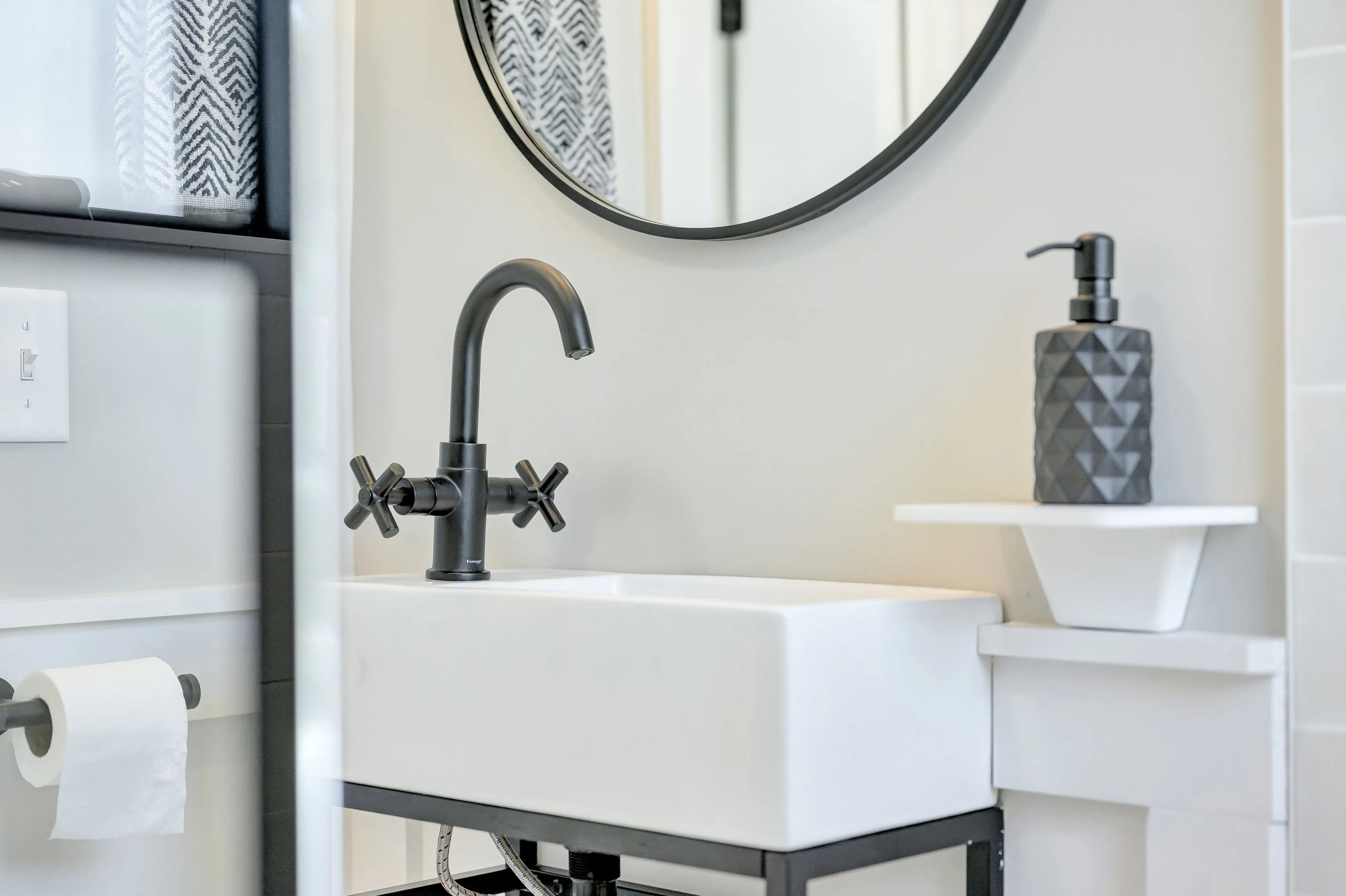Modern bathroom interior with a white rectangular vessel sink, matte black faucet, towel, round mirror, and soap dispenser.