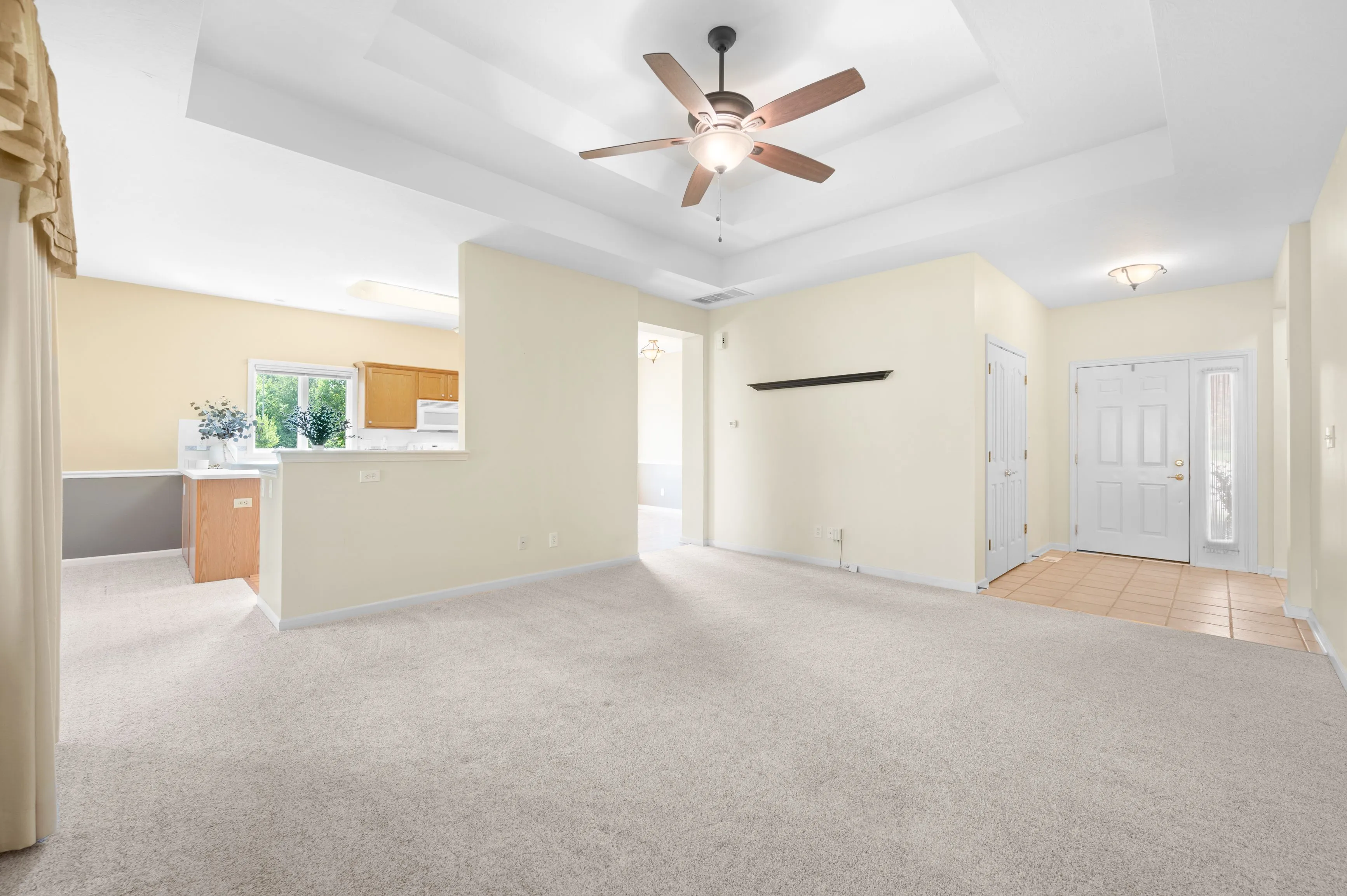 Spacious empty living room with beige walls, carpet flooring, ceiling fan, and a view into the kitchen and entryway.