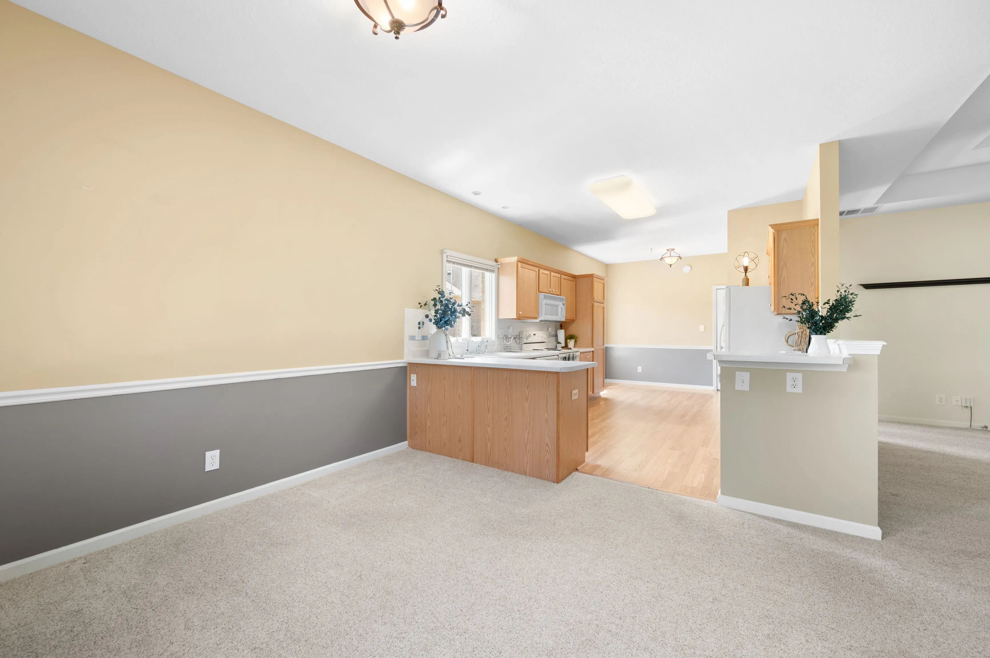Spacious and brightly lit empty room with combined kitchen and dining area, featuring wooden cabinets, carpeted and hardwood flooring, and neutral color scheme.