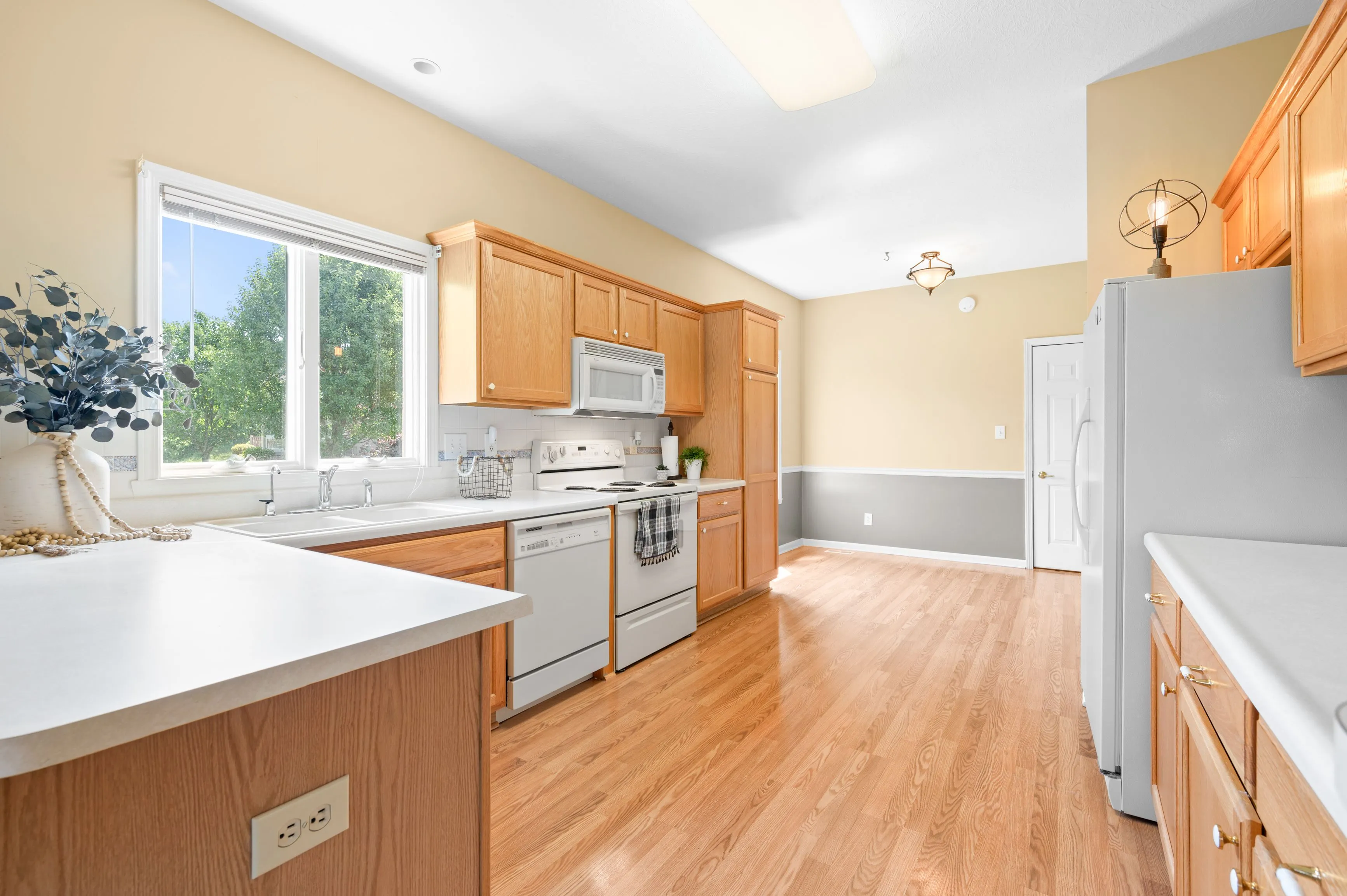 Bright and spacious kitchen interior with wooden cabinets, white appliances, hardwood flooring, and a large window with a garden view.