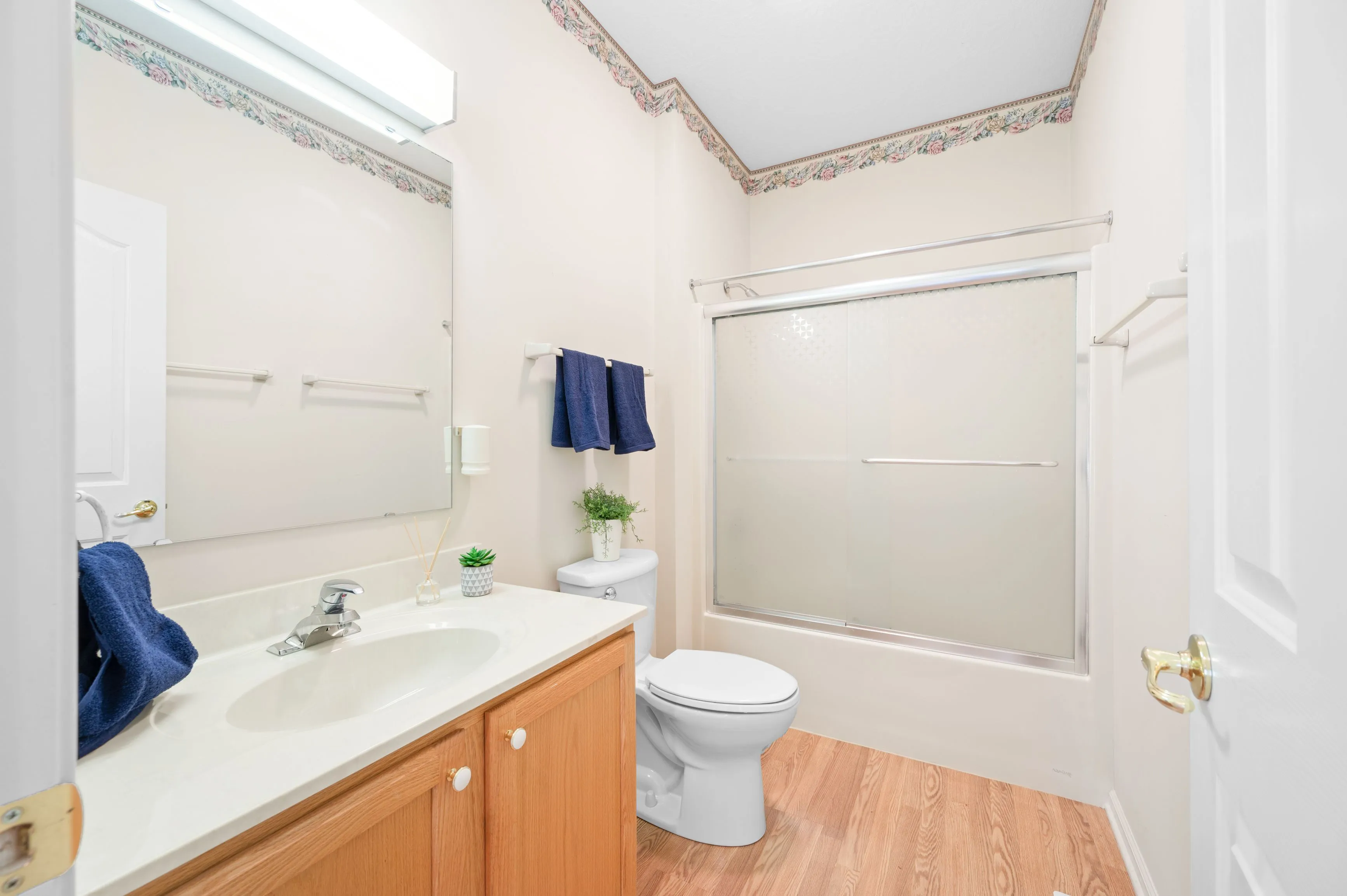 A clean and bright bathroom with wooden cabinets, a large mirror, a toilet, and a shower with a frosted glass door. Decorative elements include blue towels and small potted plants.