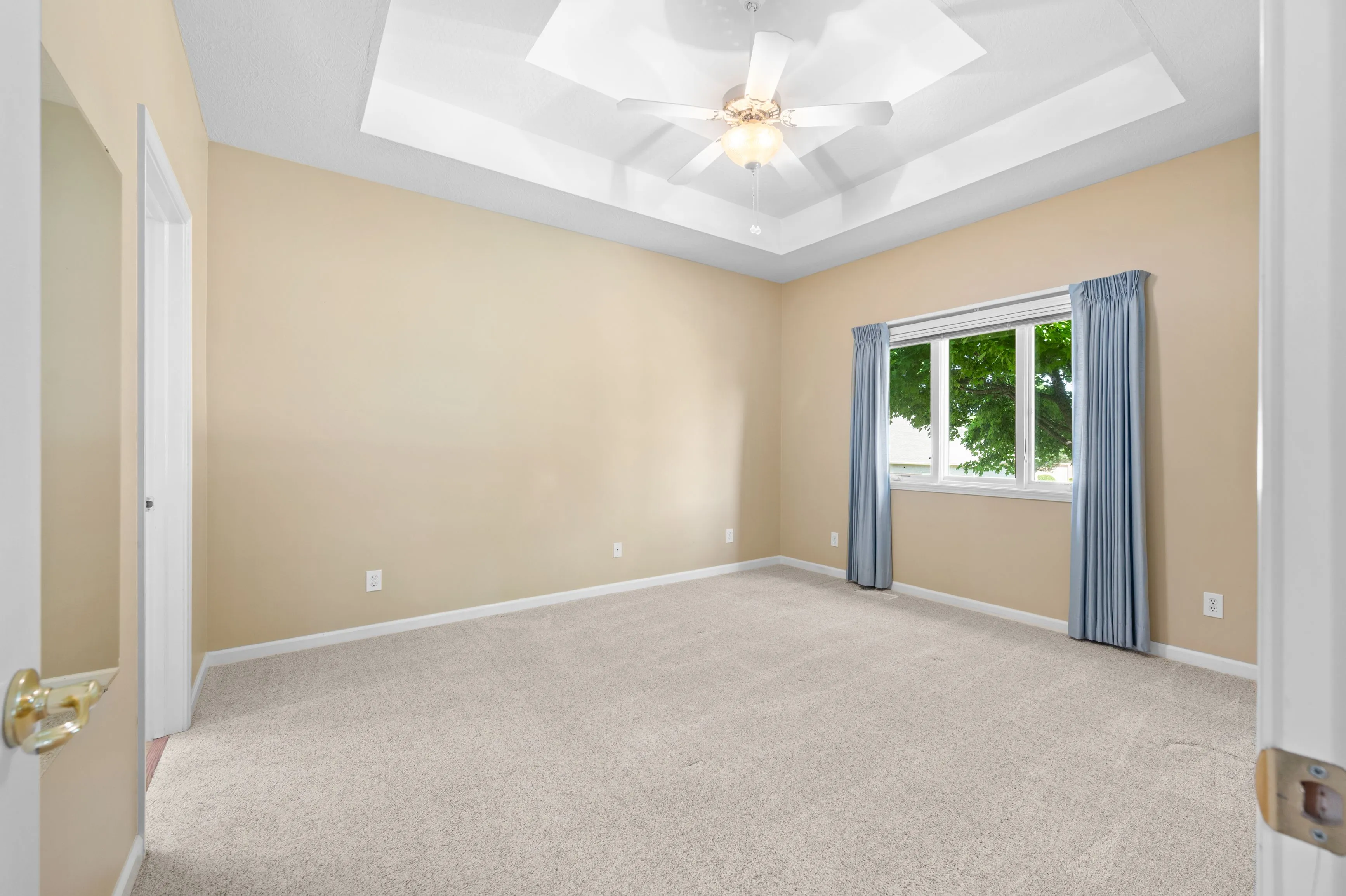 Empty room with beige walls, carpet flooring, window with blue curtains, and a ceiling fan.