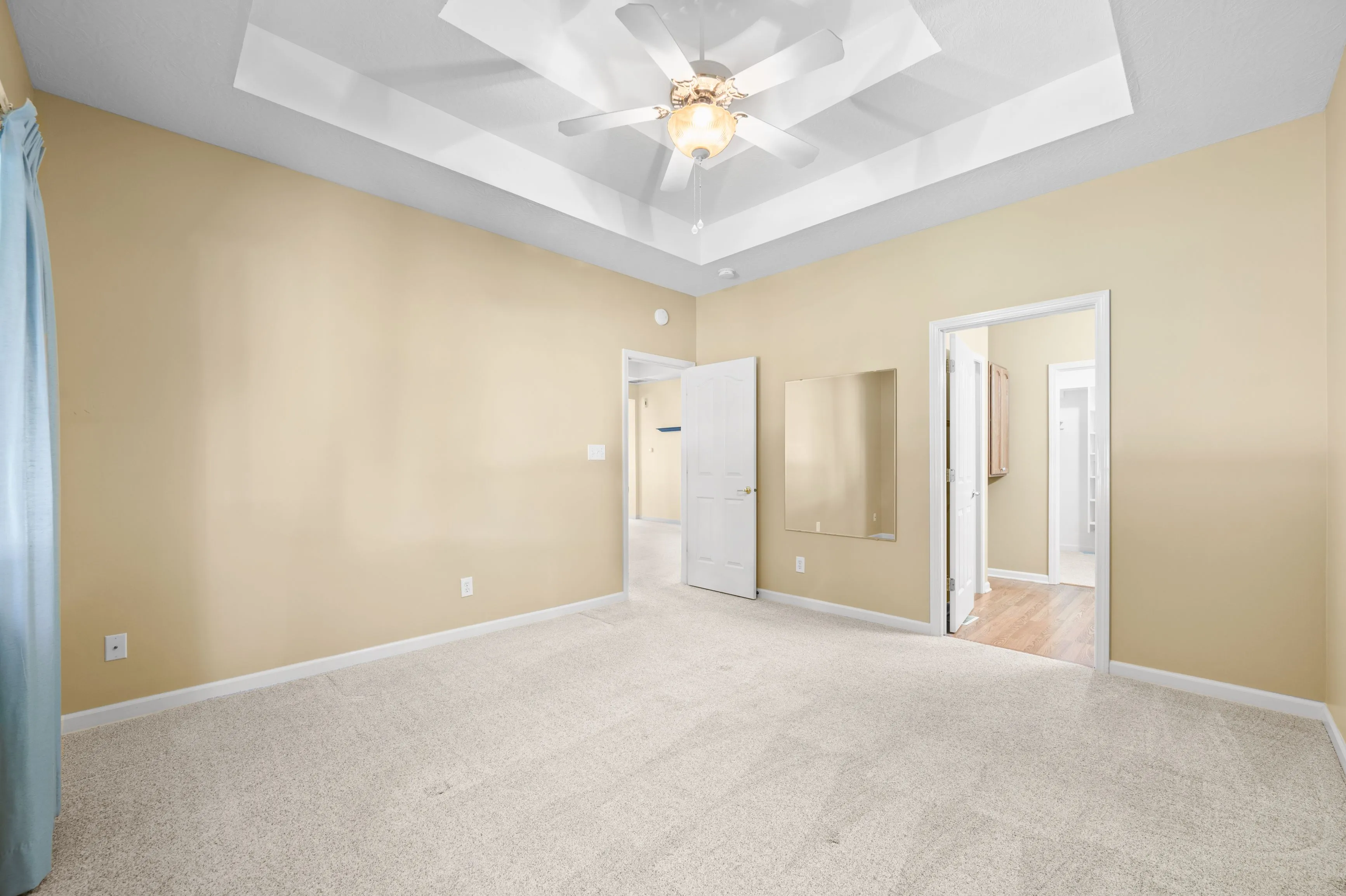 Empty room with beige walls, white ceiling fan, and carpeted floor, showing open doors to adjacent rooms.