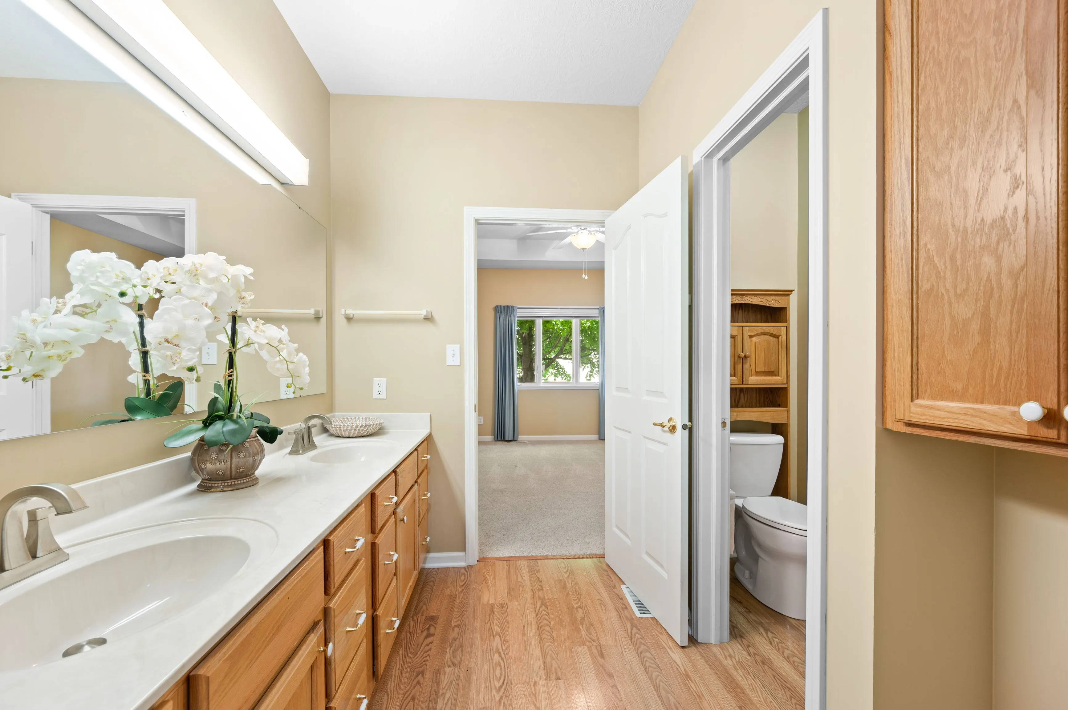 Bright bathroom interior featuring a long vanity with a white countertop, double sinks, a large mirror, and a wooden cabinet, with a toilet visible in a separate room through an open door. A bouquet of white orchids adds a decorative touch, and a doorway to an adjoining room with a window suggests a suite layout.