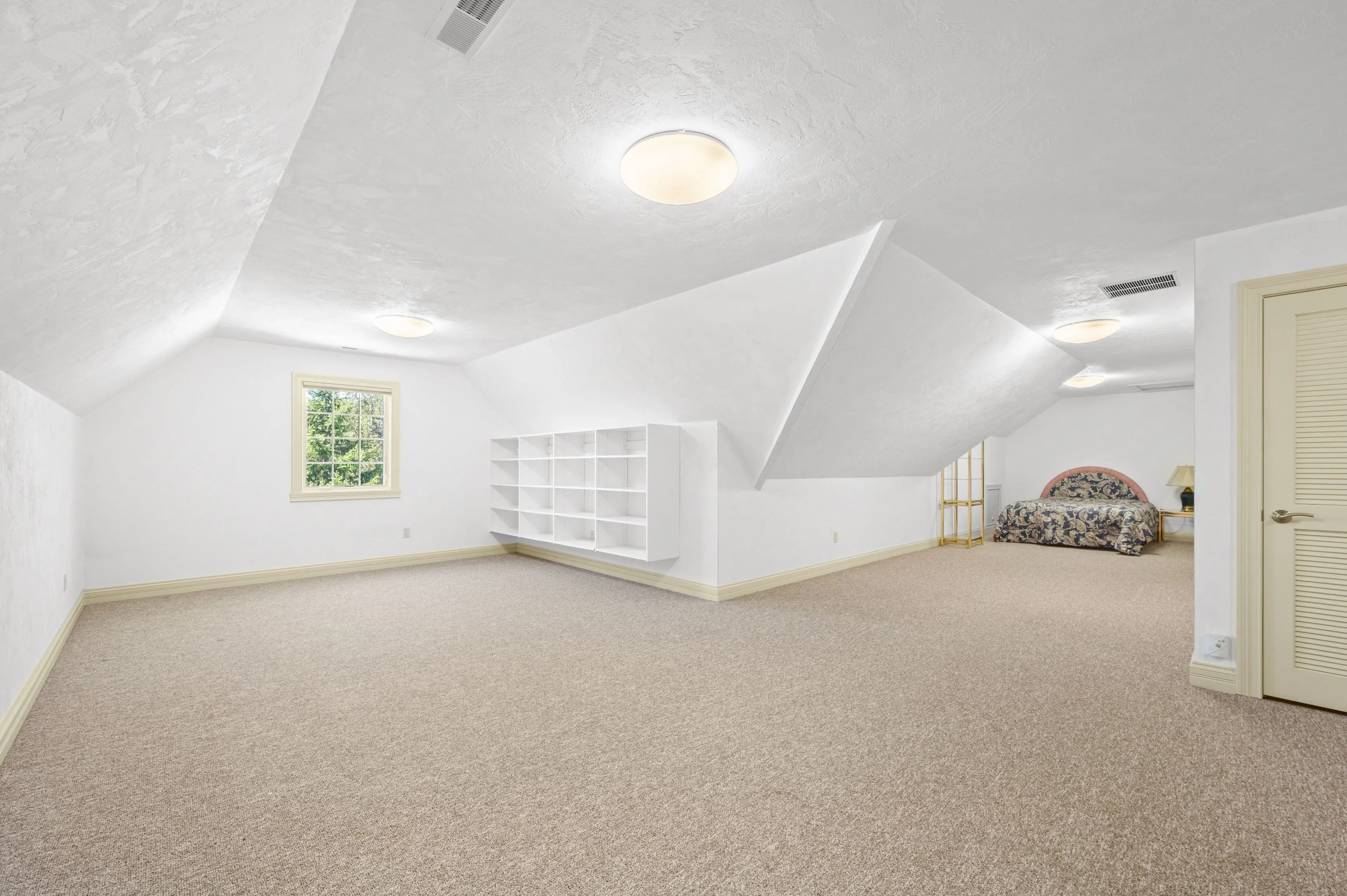 Bright and spacious attic room with slanted ceilings, carpeted floor, white walls, built-in shelving unit, and overhead lighting, leading to an adjoining room with a bed visible in the background.