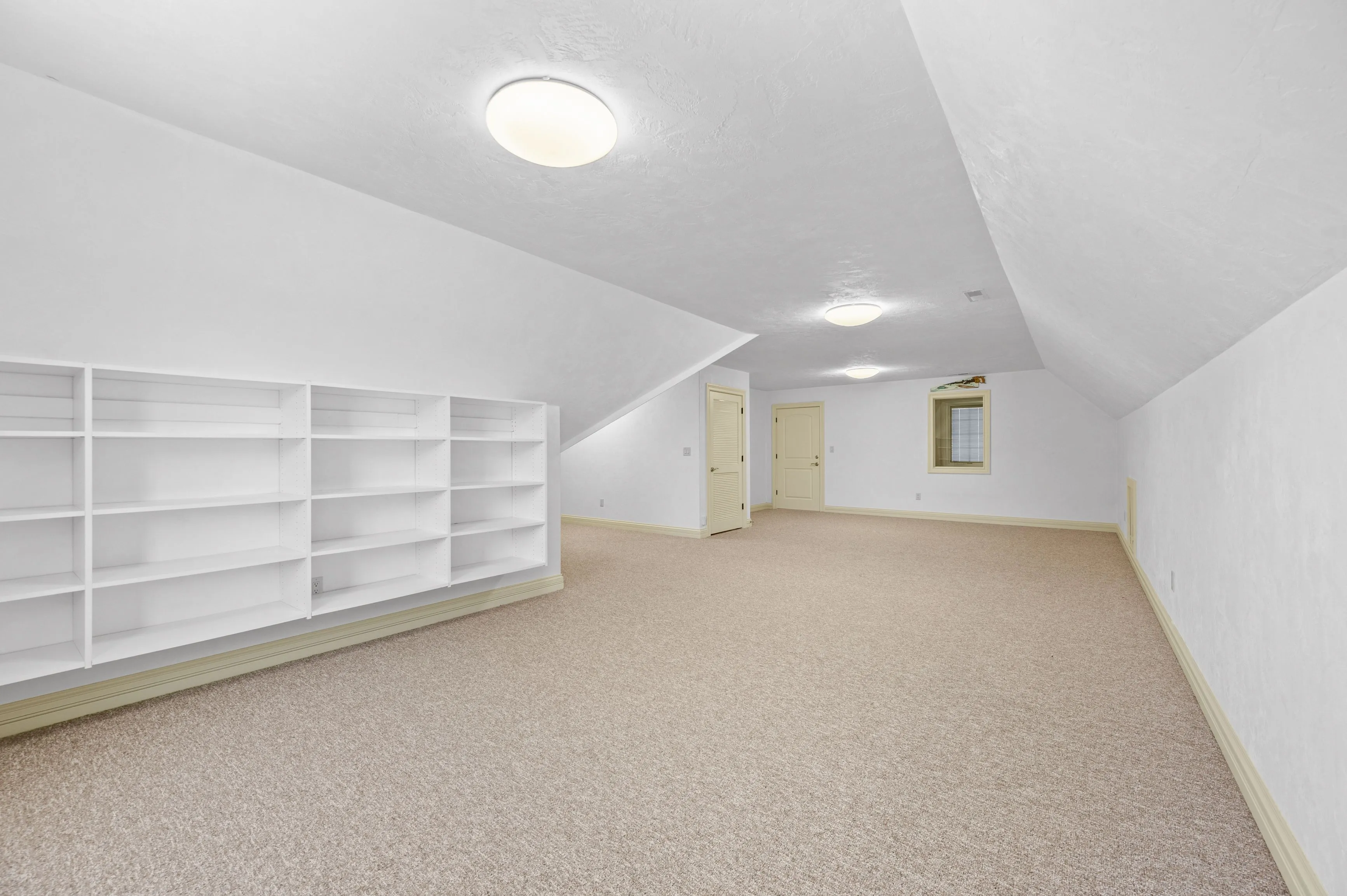 Bright, empty attic room with beige carpet, white walls, slanted ceilings, two ceiling lights, and a built-in white bookshelf.