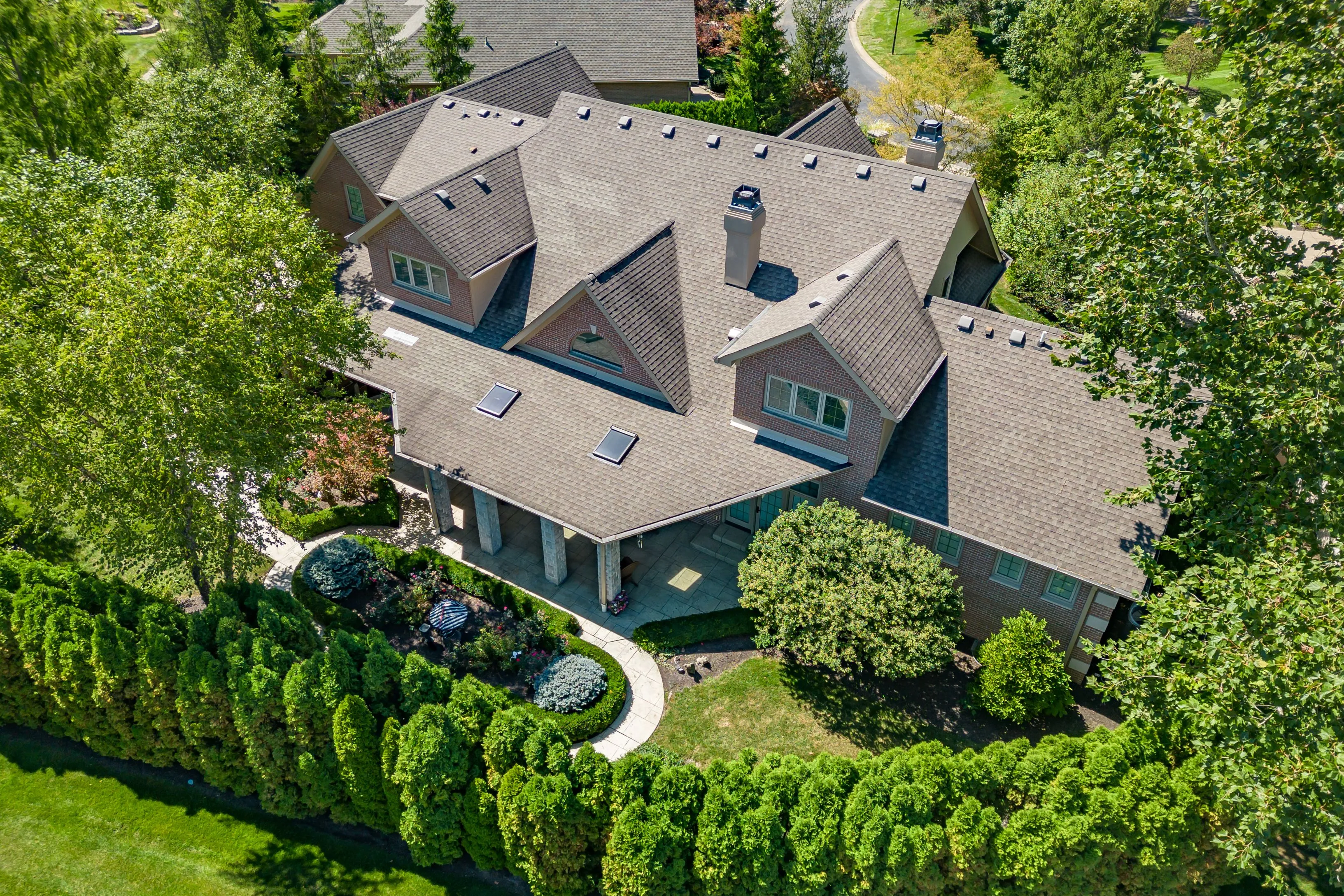 Aerial view of a large suburban house with a multi-gabled roof, surrounded by a landscaped garden and a hedge perimeter.