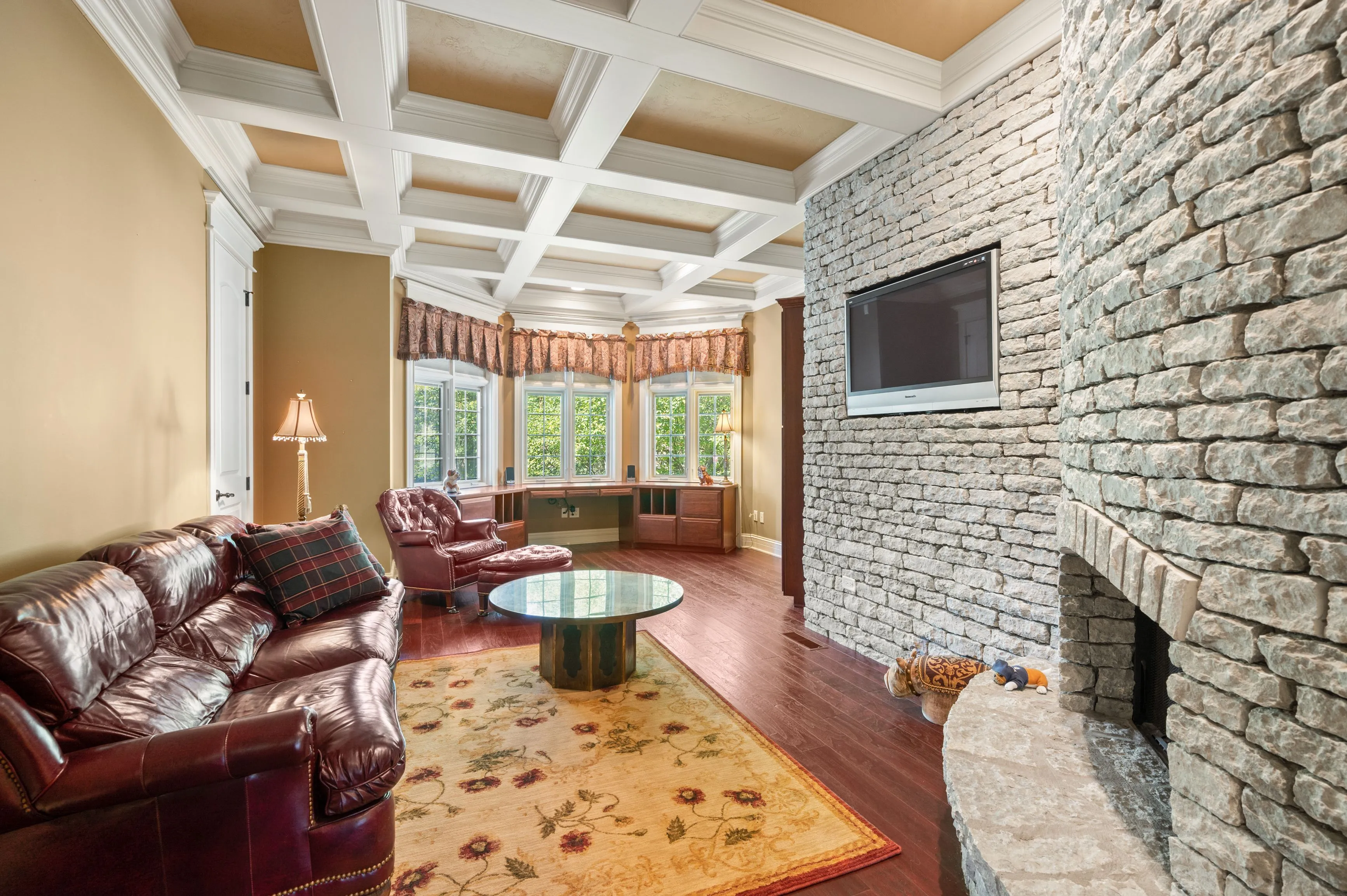 Elegant living room with coffered ceiling, stone fireplace, leather sofas, hardwood floors, and a bay window.