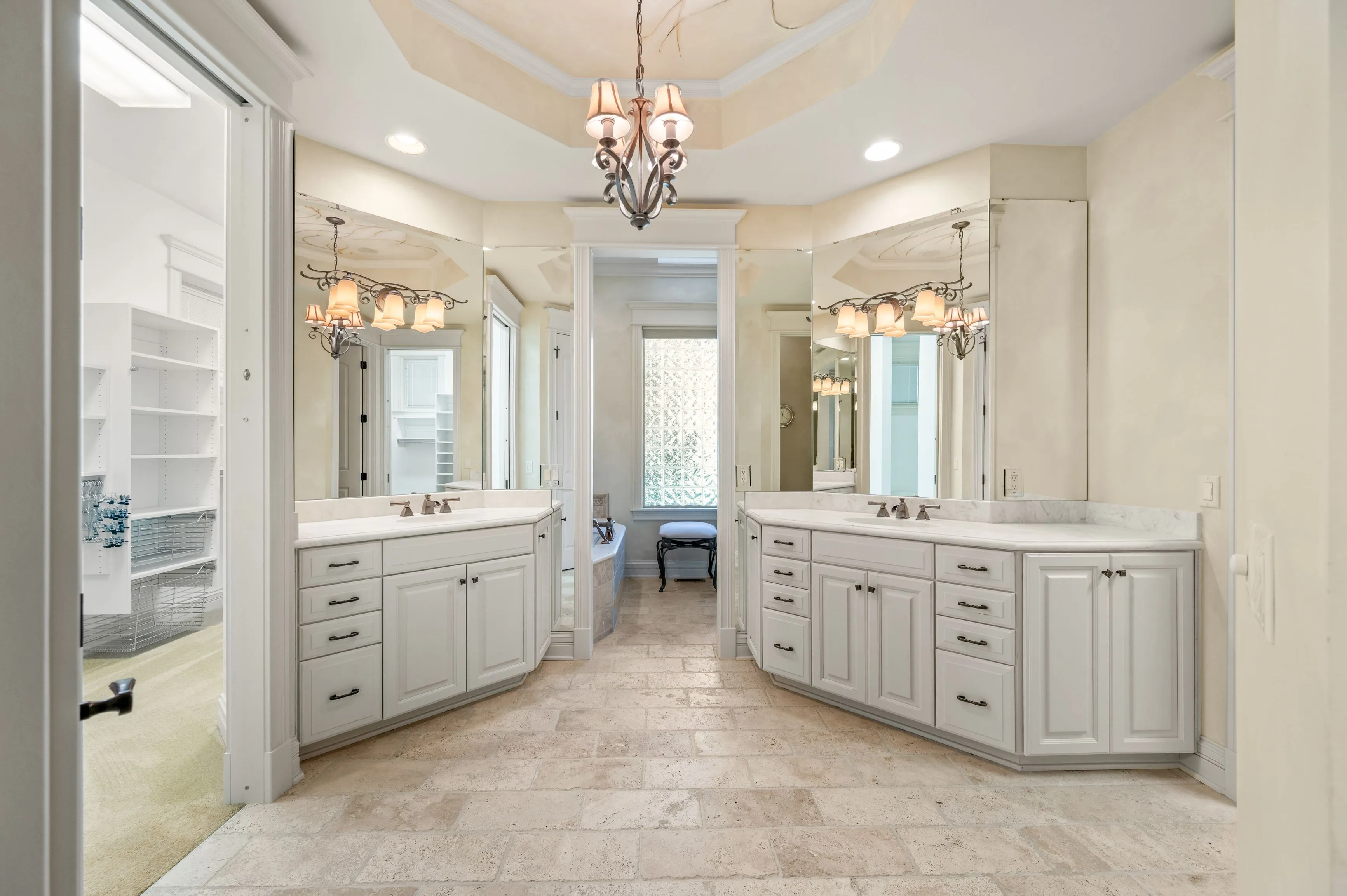 Elegant, spacious bathroom interior with dual white vanities, marble countertops, and a stone-tiled floor, leading to a frosted glass window and a separate shower area.