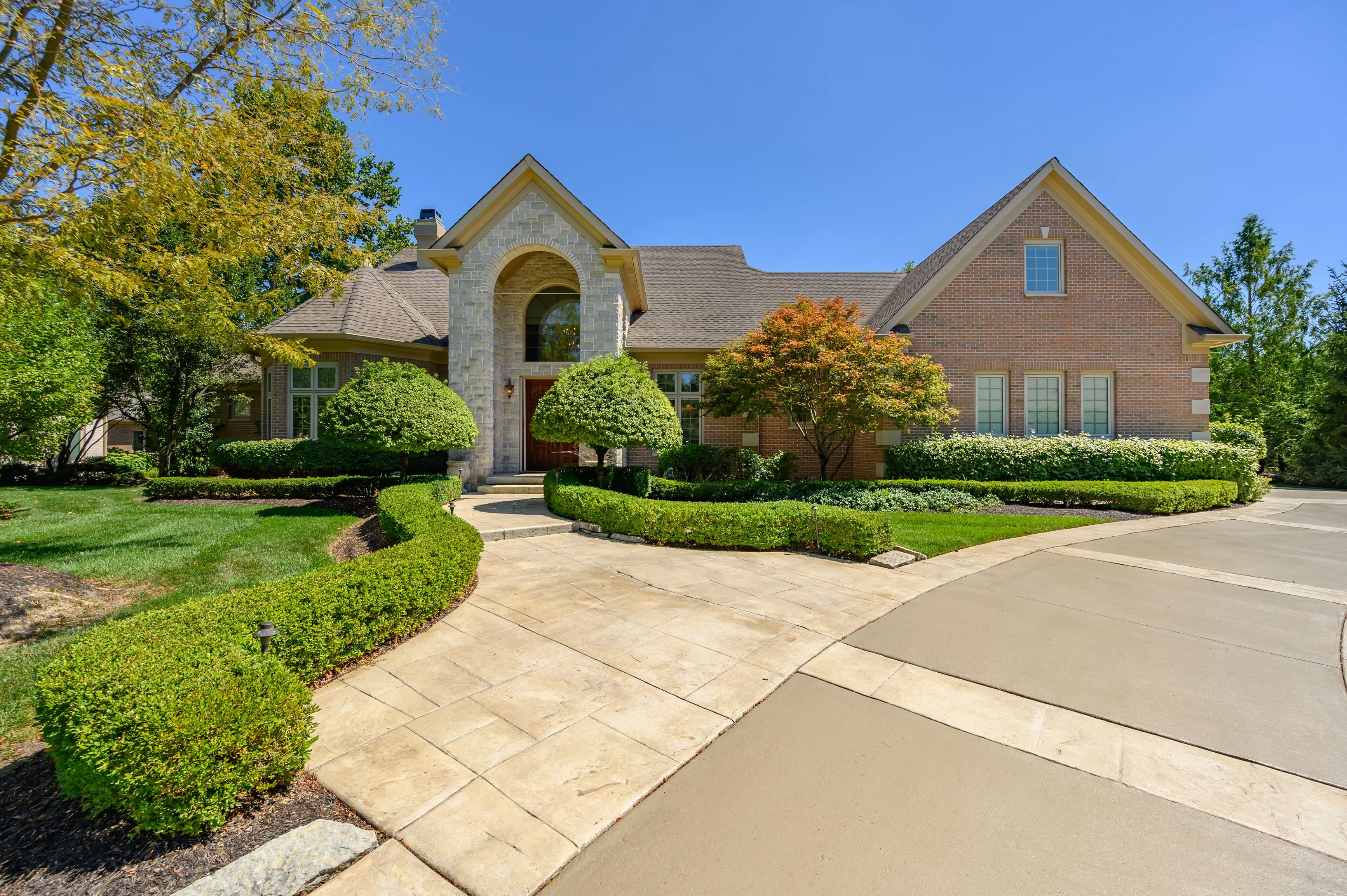 Elegant two-story brick house with an arched entryway, manicured landscaping, and a stone walkway under a clear blue sky.