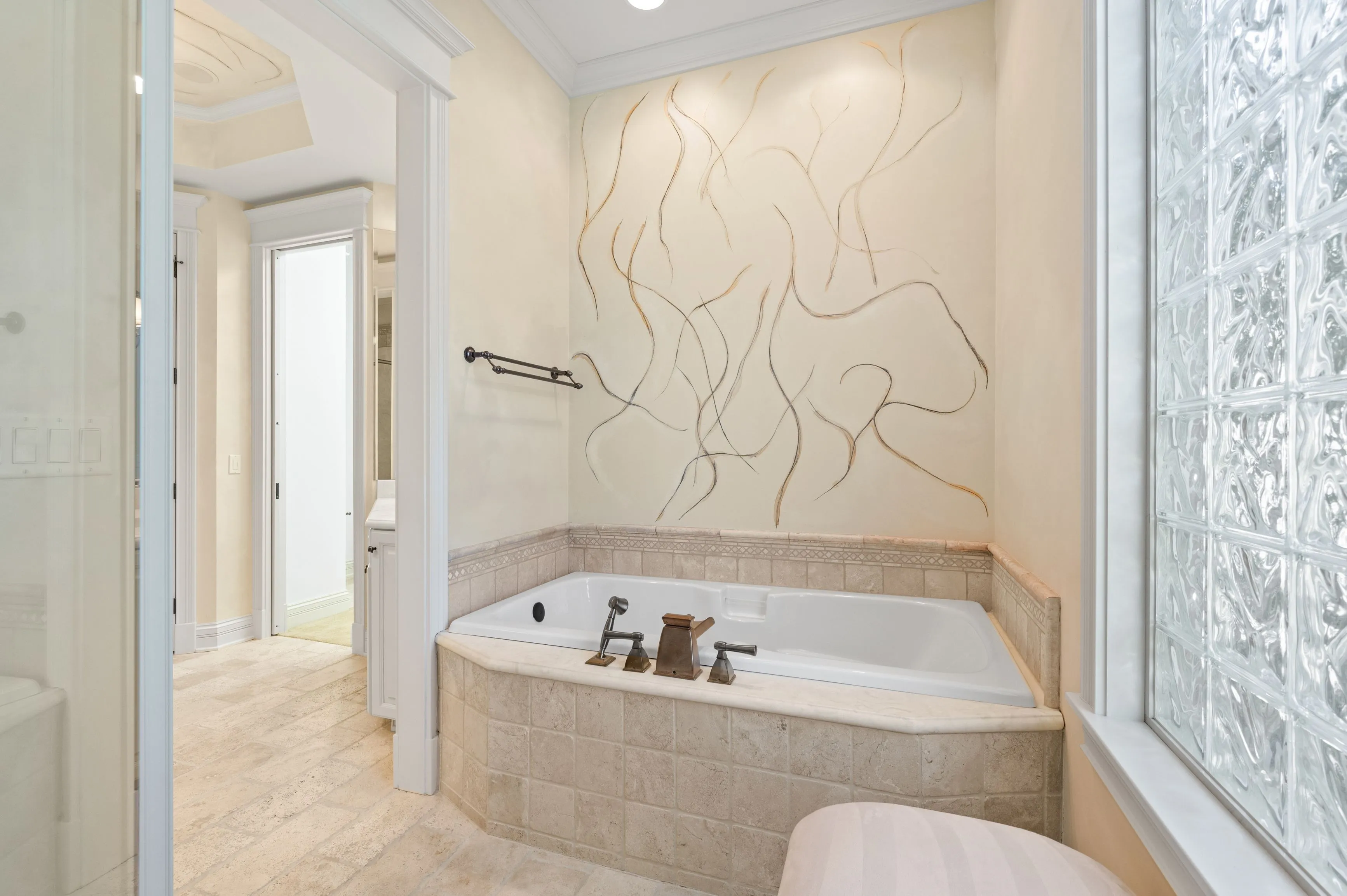 Elegant bathroom interior featuring a corner bathtub with decorative beige tiles, an artistic wall mural, glass block window, and a separate shower area with glass door.