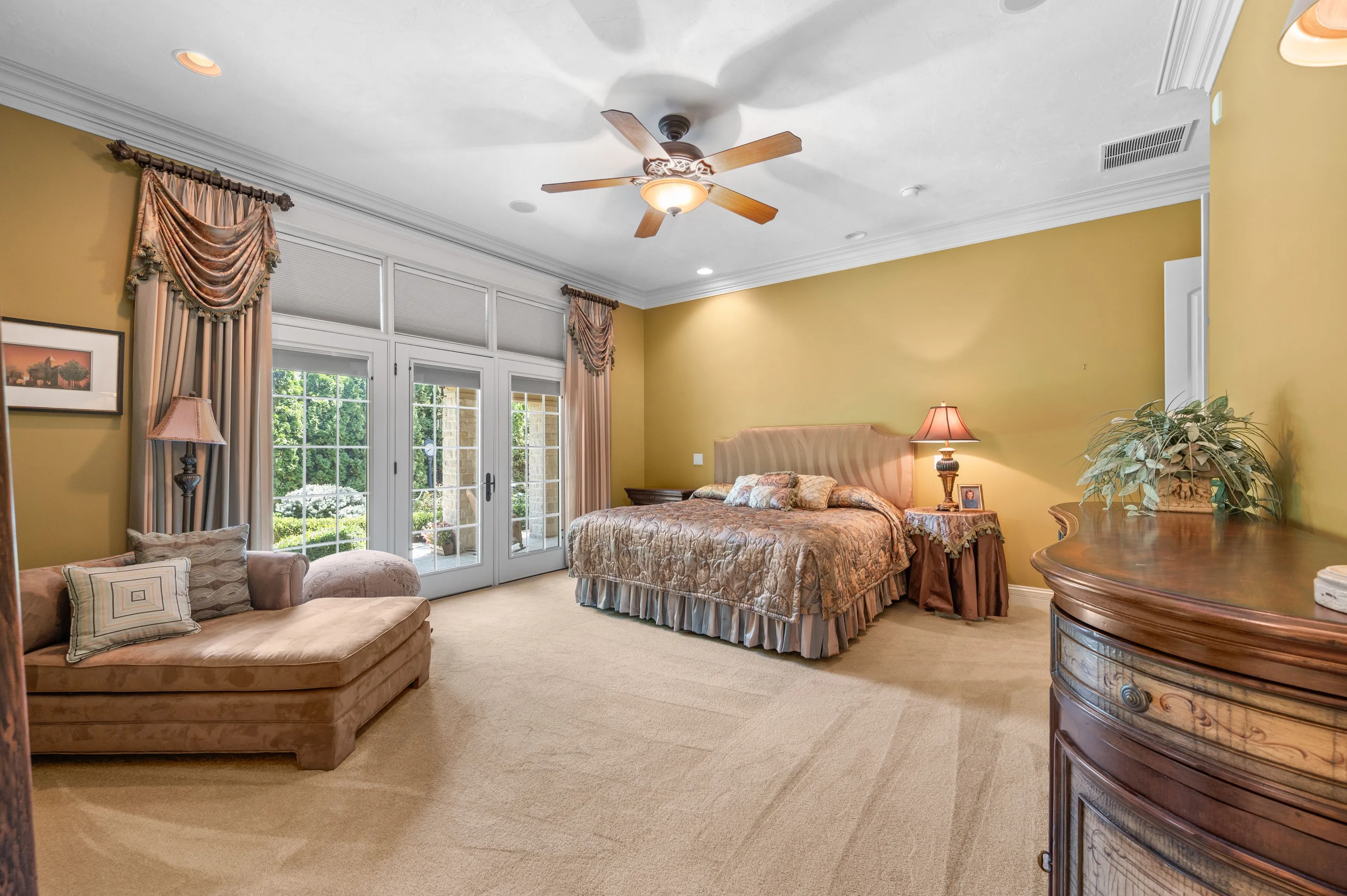 Spacious bedroom with yellow walls, a large bed with patterned bedding, elegant drapes on windows, ceiling fan, and plush carpeting.