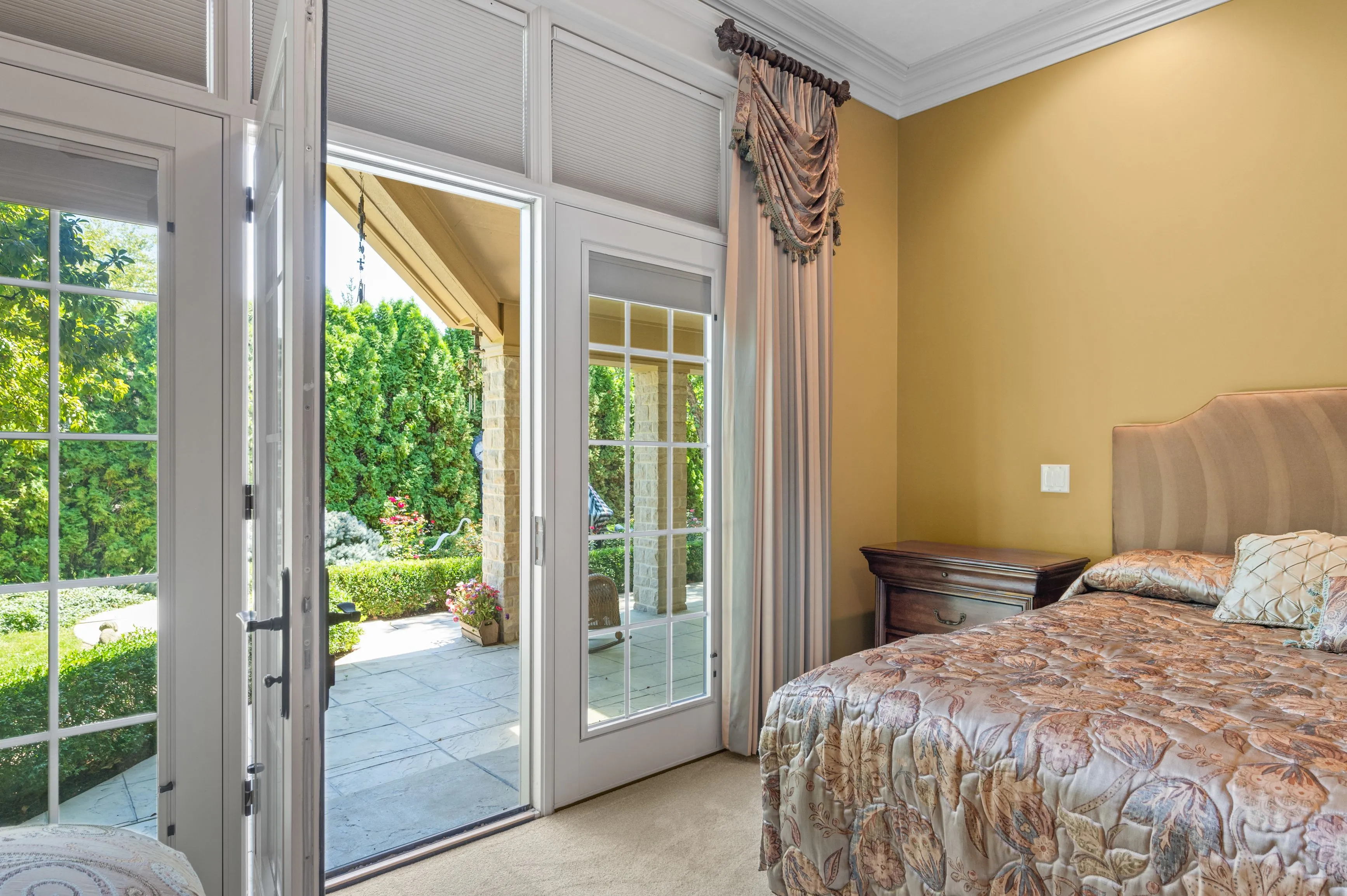 Bright bedroom with an open French door leading to a garden patio, elegant drapery, and a floral-patterned bedspread.