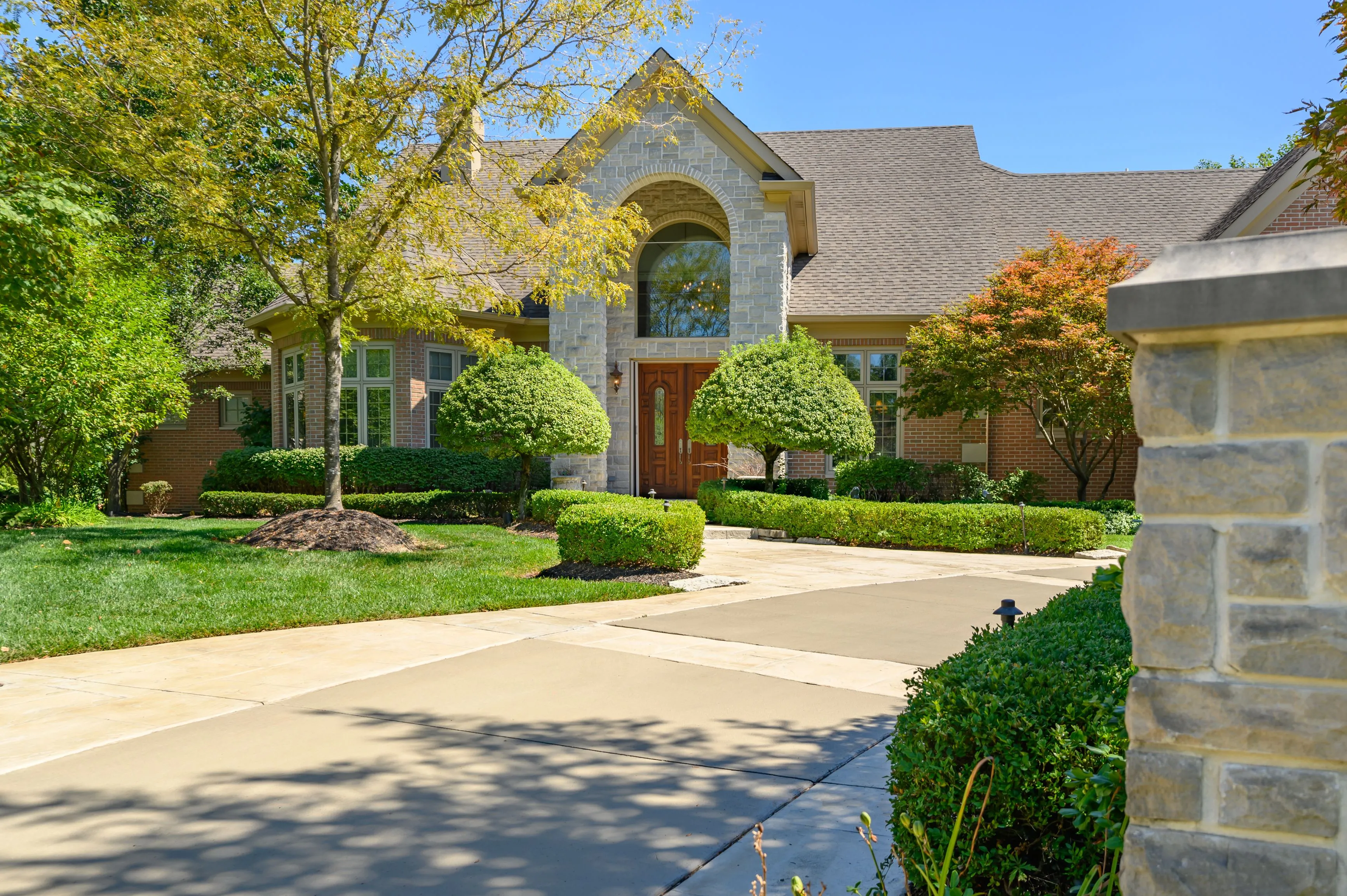 A beautiful suburban home with a landscaped front yard, featuring a stone arched entryway, pruned shrubs, and a well-maintained lawn under a blue sky.