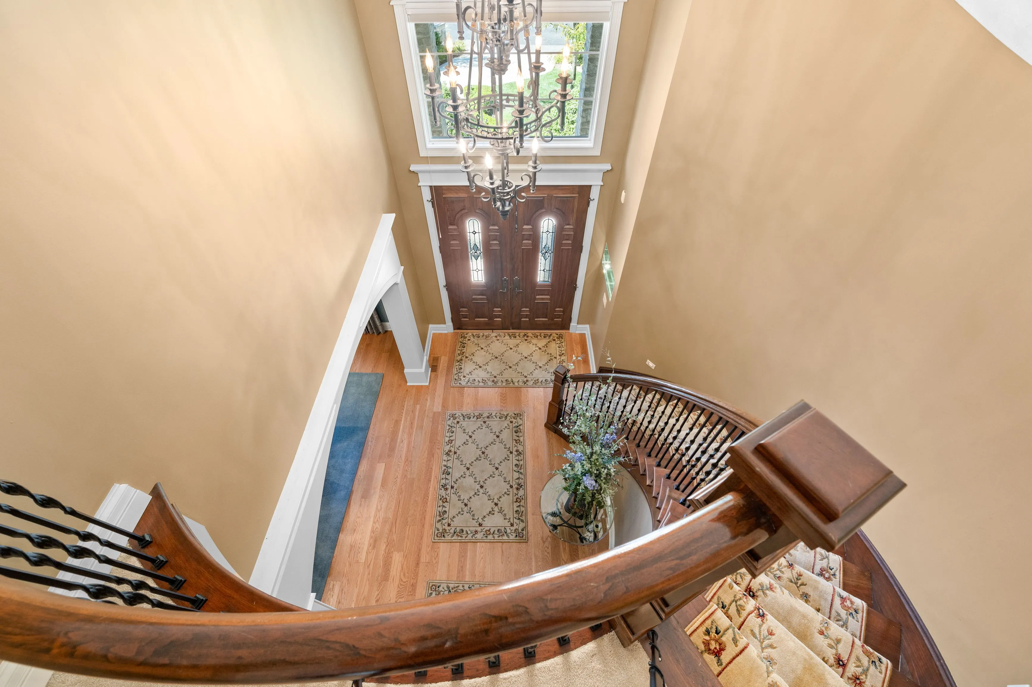 Elegant home entryway seen from the top of a staircase with hardwood floors, a large chandelier, and a double front door with glass panels.