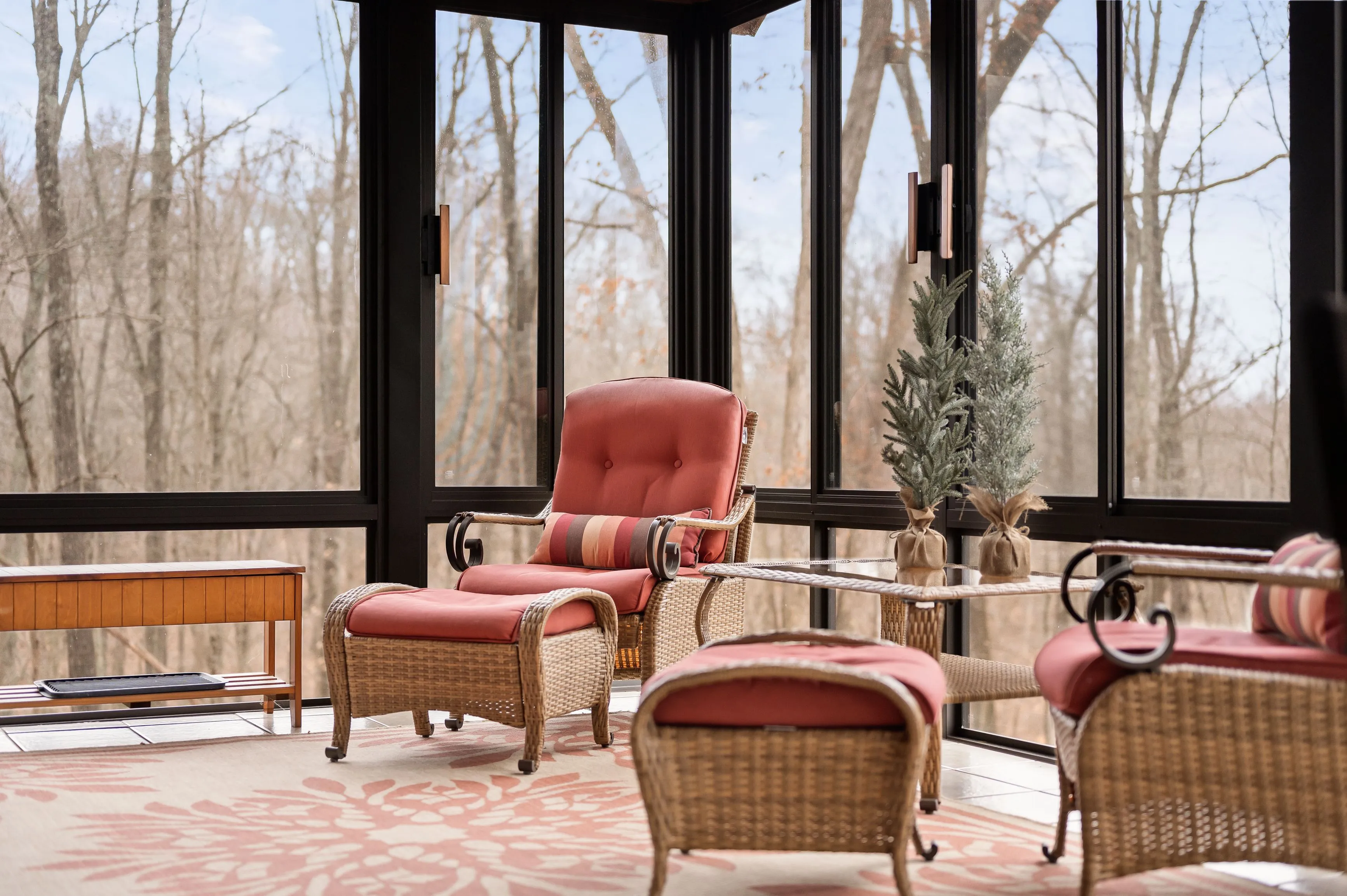 Cozy sunroom with wicker furniture, a red cushioned armchair and ottoman, and large windows overlooking bare trees.