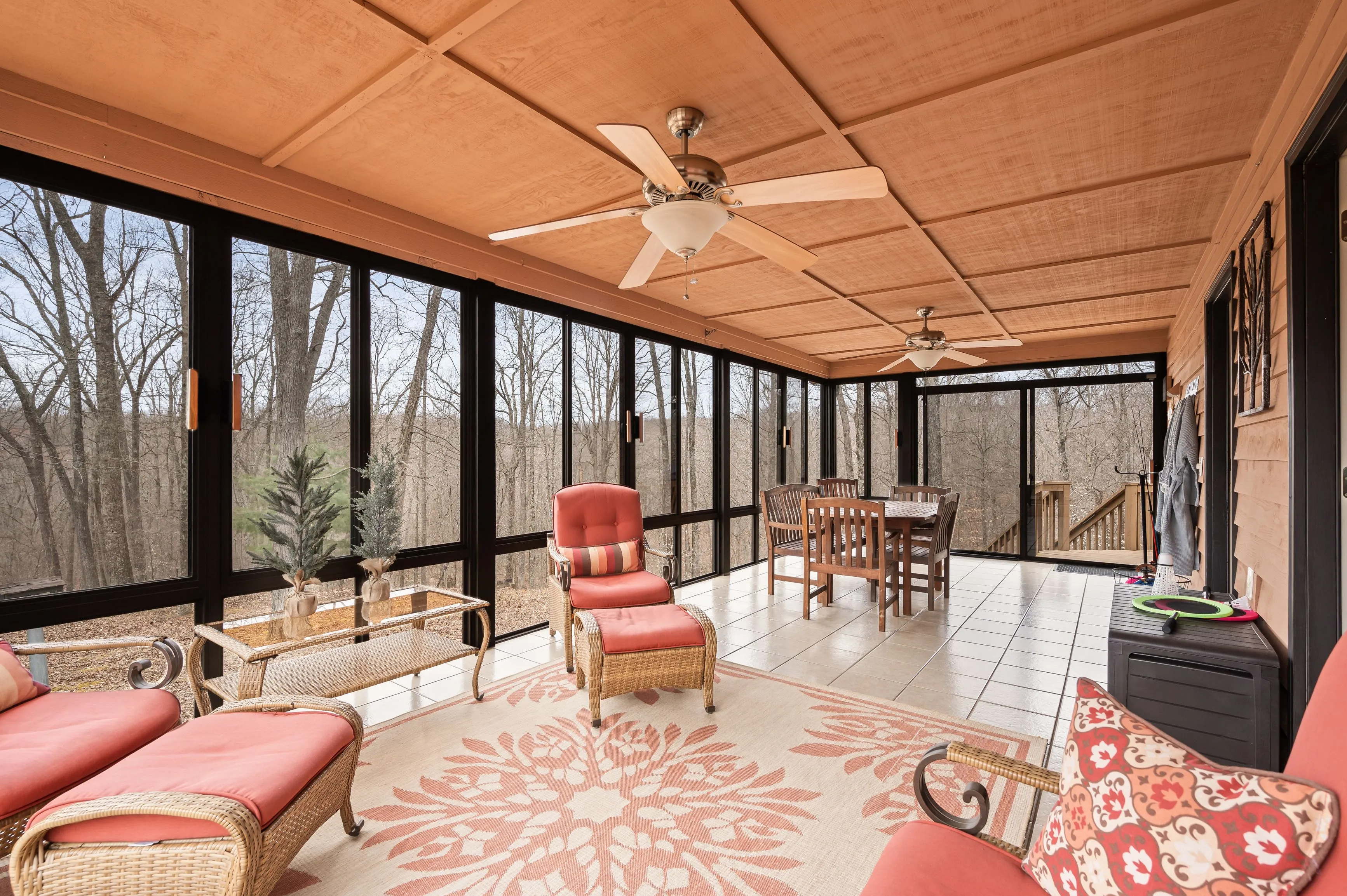 Spacious screened porch with wooden furniture, red cushions, ceiling fans, and a view of leafless trees.