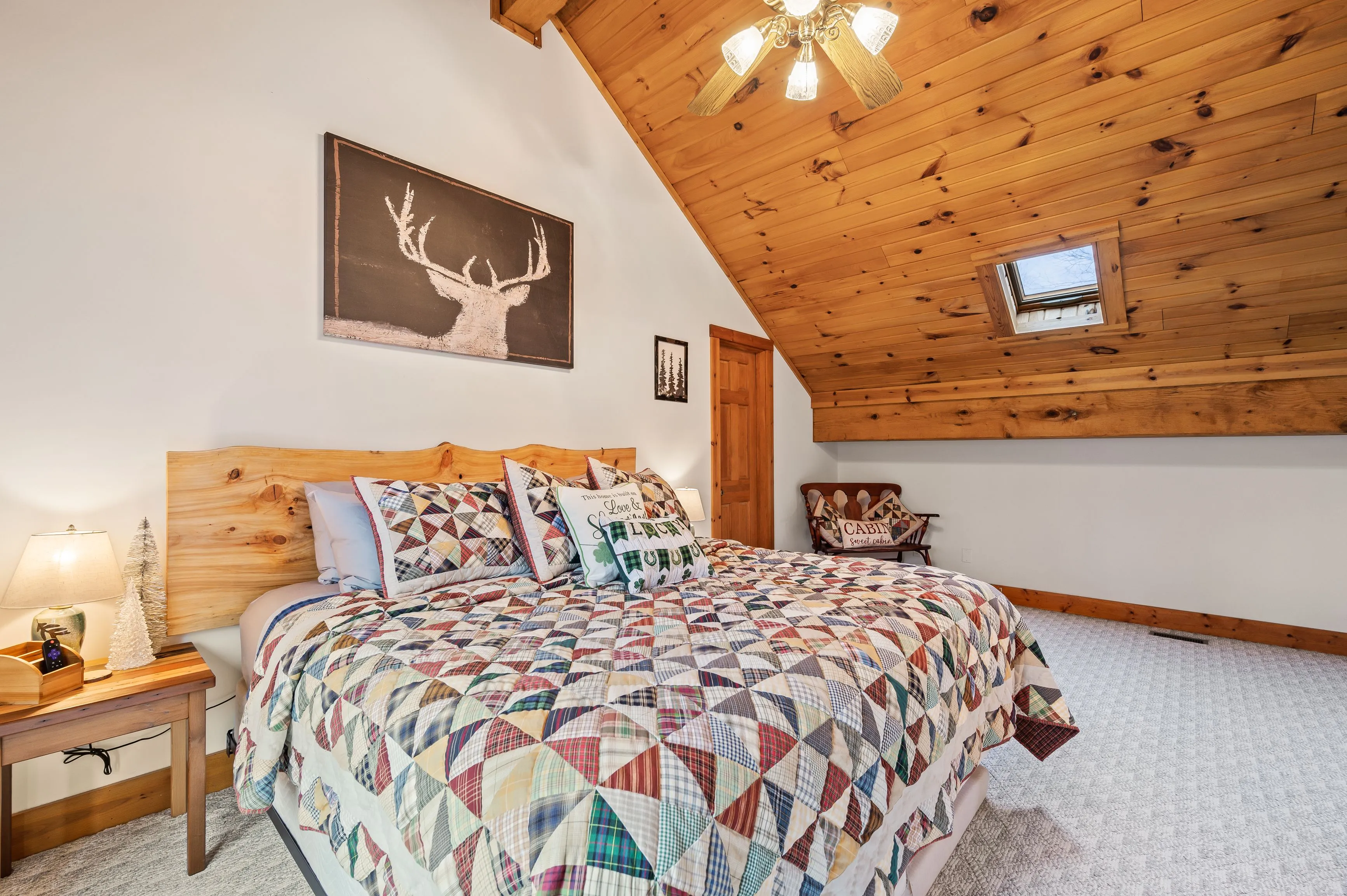 Cozy cabin-style bedroom with wooden ceiling and walls, quilt-covered bed, and rustic decor including a deer head painting.