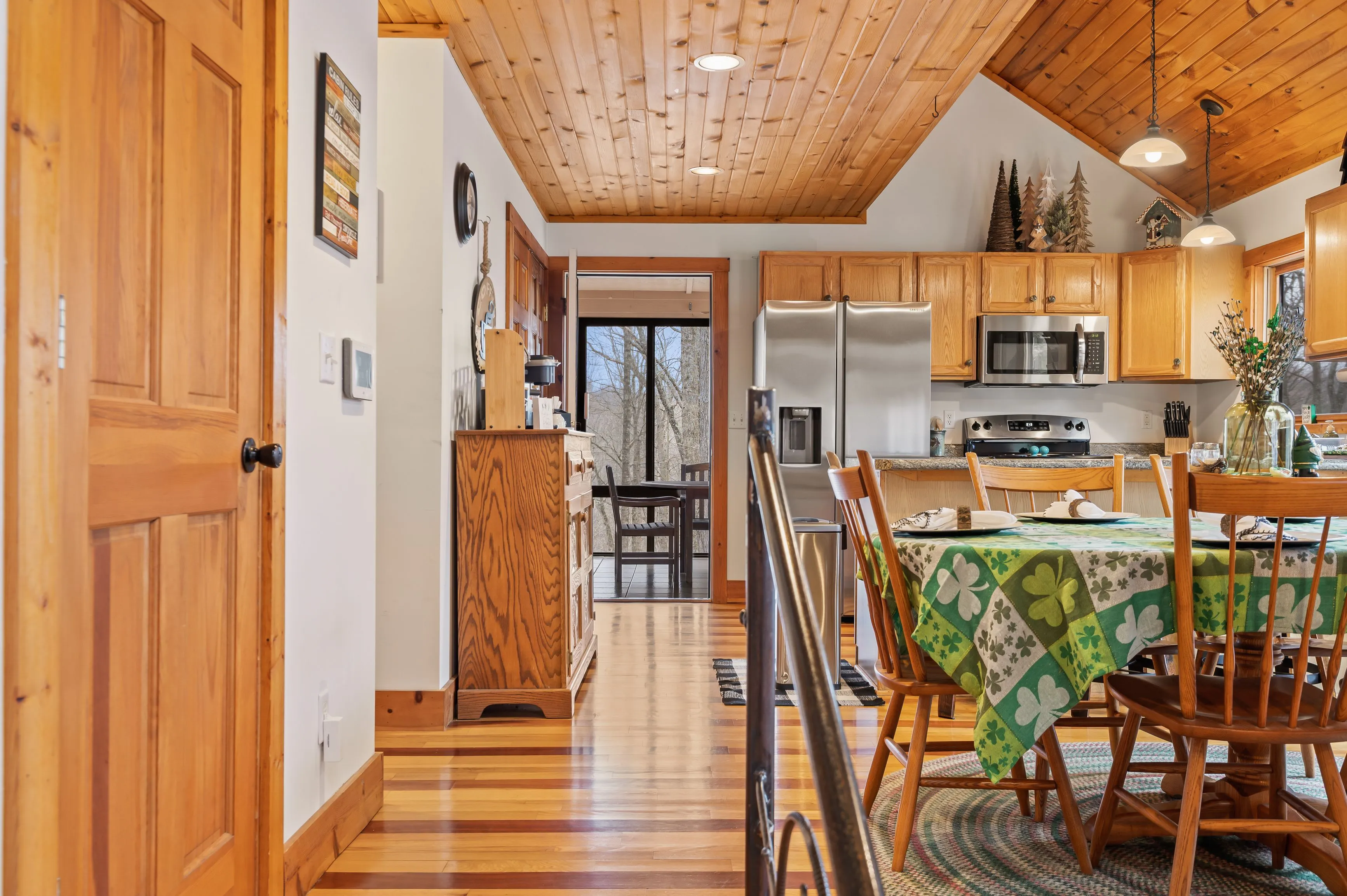 Cozy wooden cabin interior with open concept kitchen and dining area, featuring stainless steel appliances, a wooden dining table with a clover-patterned tablecloth, and view to a deck area through sliding doors.