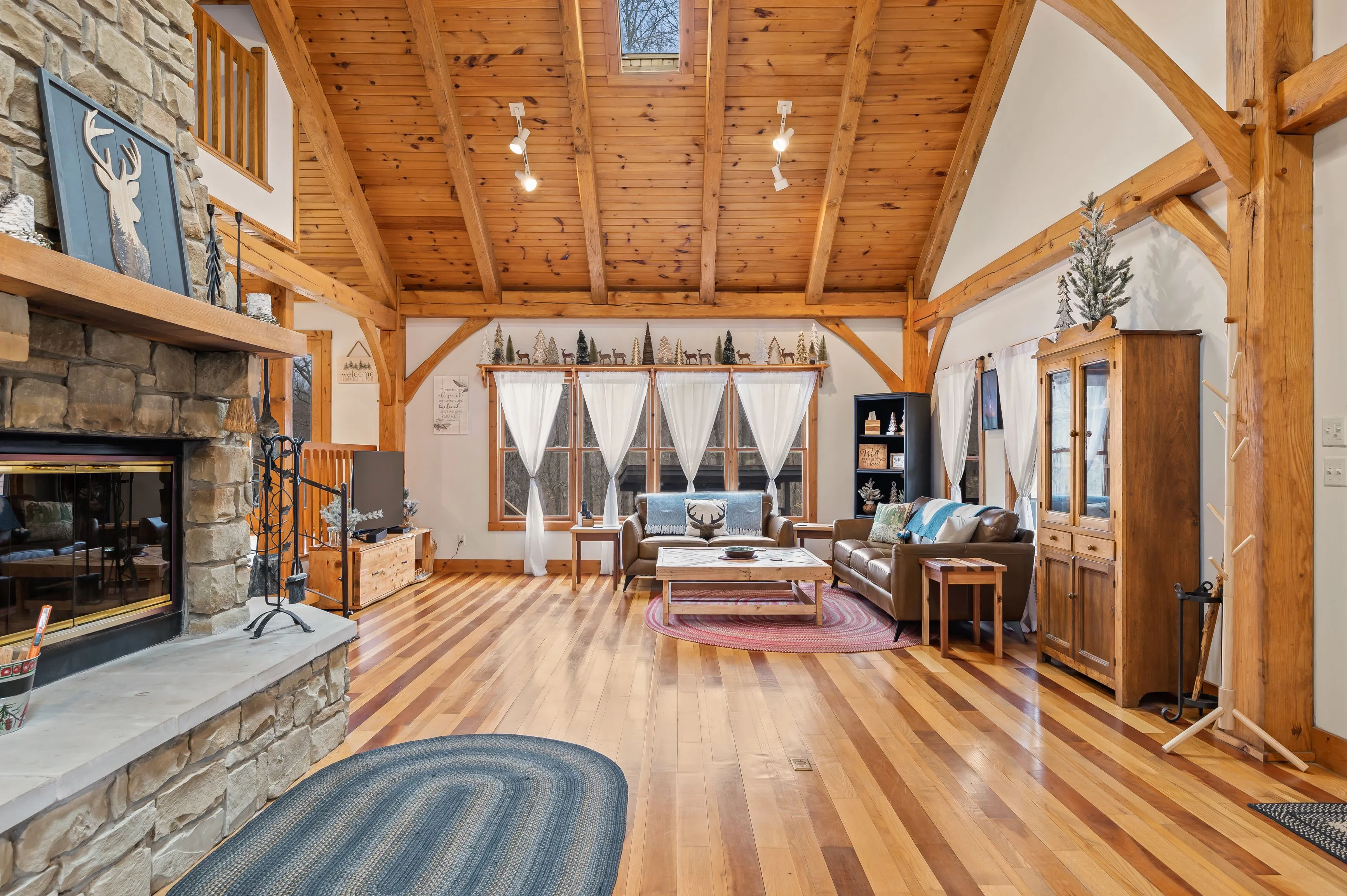 Spacious living room interior with high wooden ceilings, exposed beams, hardwood floors, stone fireplace, and large windows draped with sheer curtains in a rustic cabin style home.