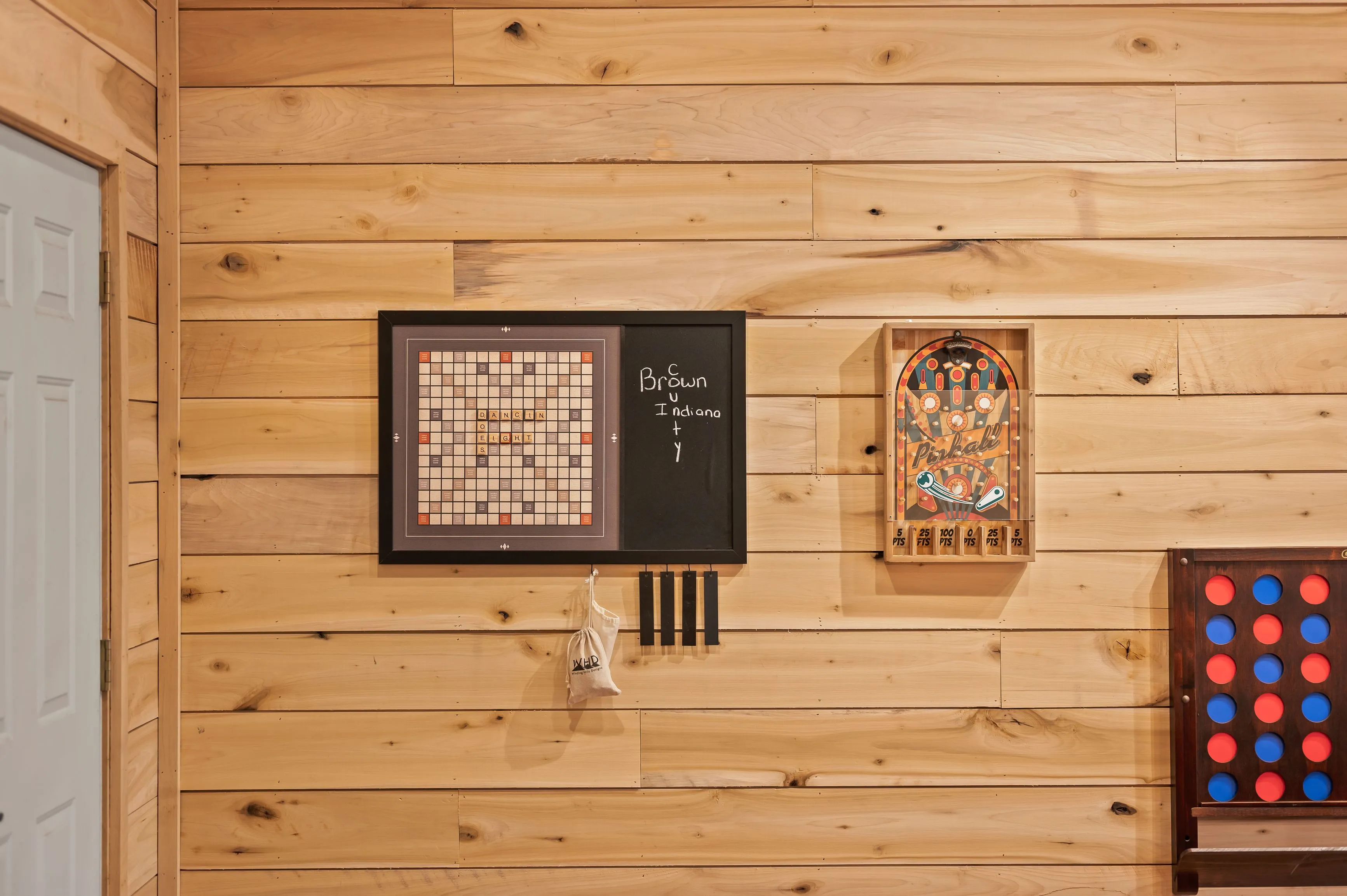 Wood-paneled room with a framed Scrabble board, a chalkboard with writing, a decorative pinball backglass, and a Connect Four game on the wall.