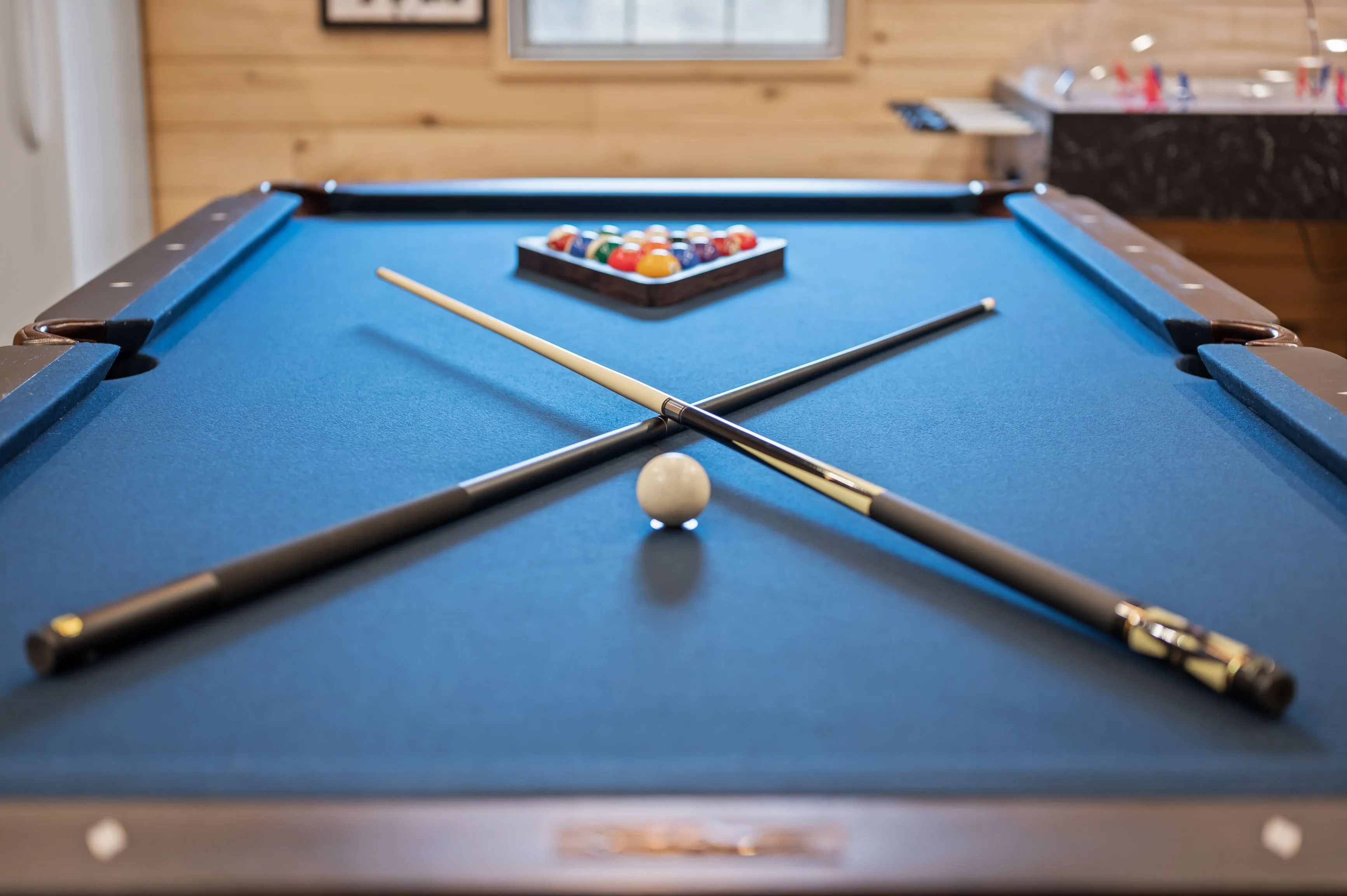 Pool cues crossed over a white ball on a blue billiard table with a rack of colorful billiard balls in the background.