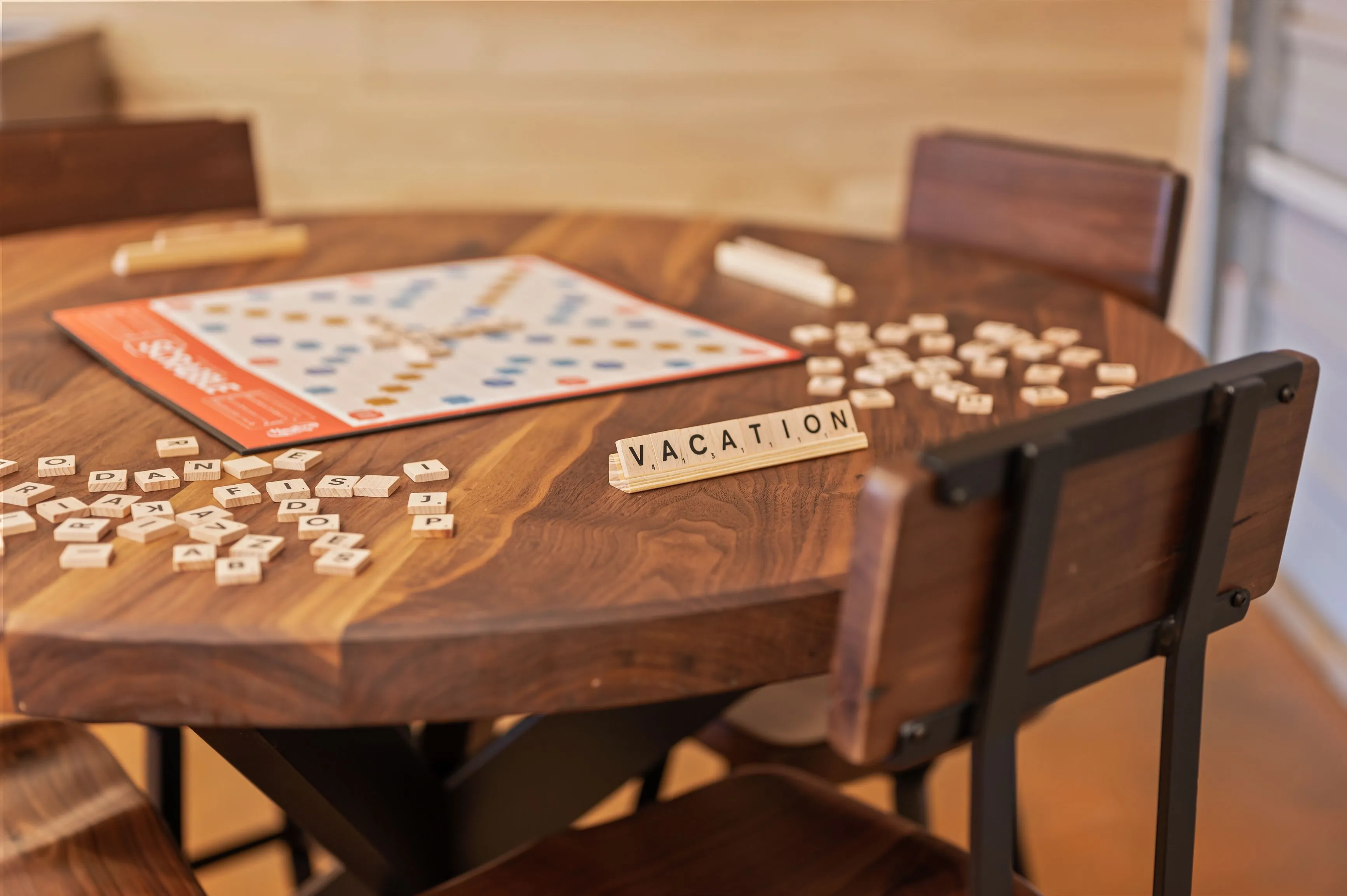Wooden table with a Scrabble board game and tiles, a rack with the letters spelling "VACATION," and chairs around the table.