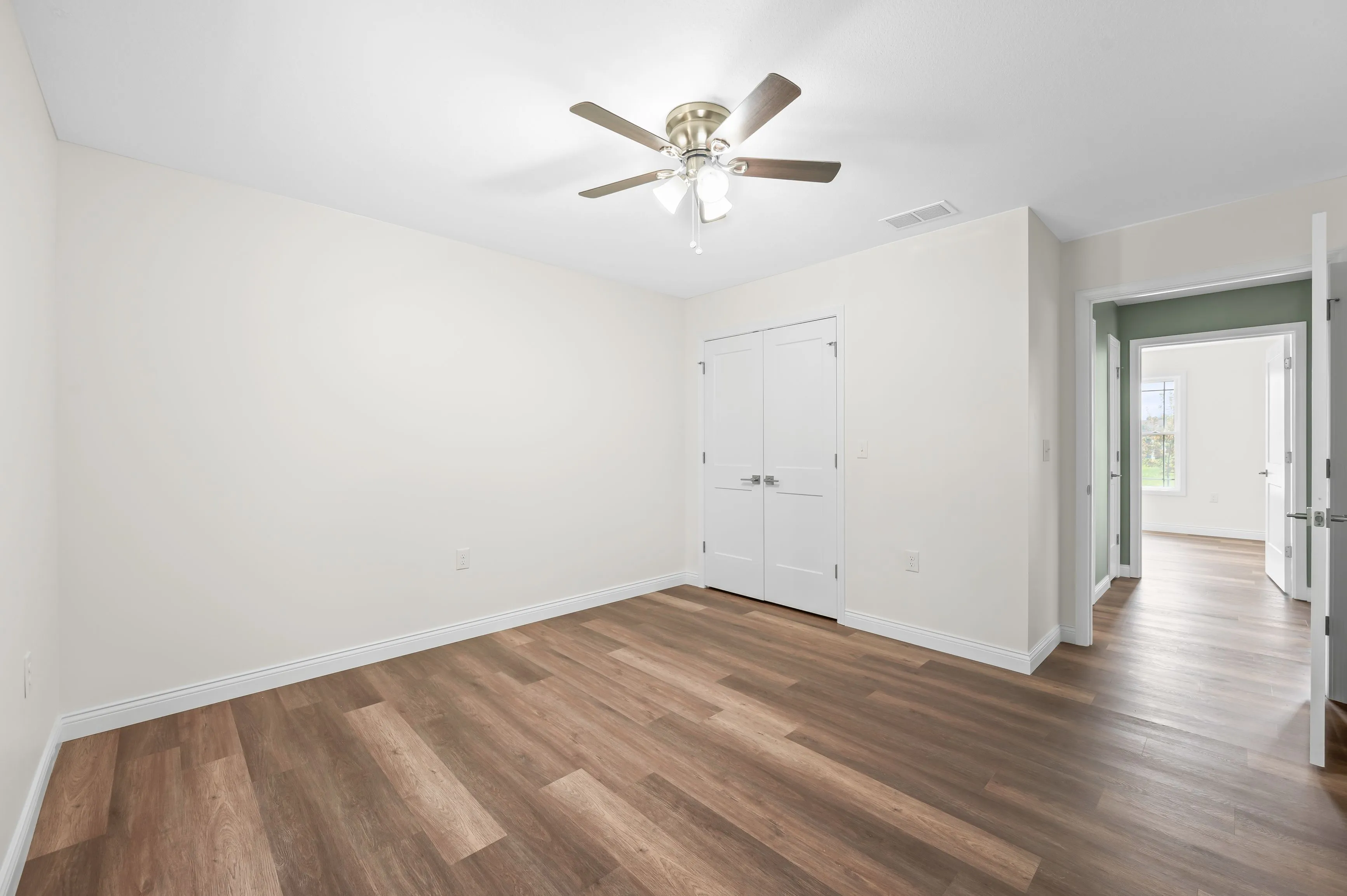 Bright, empty room with white walls, wood flooring, a ceiling fan with lights, and open doors leading to adjacent rooms.