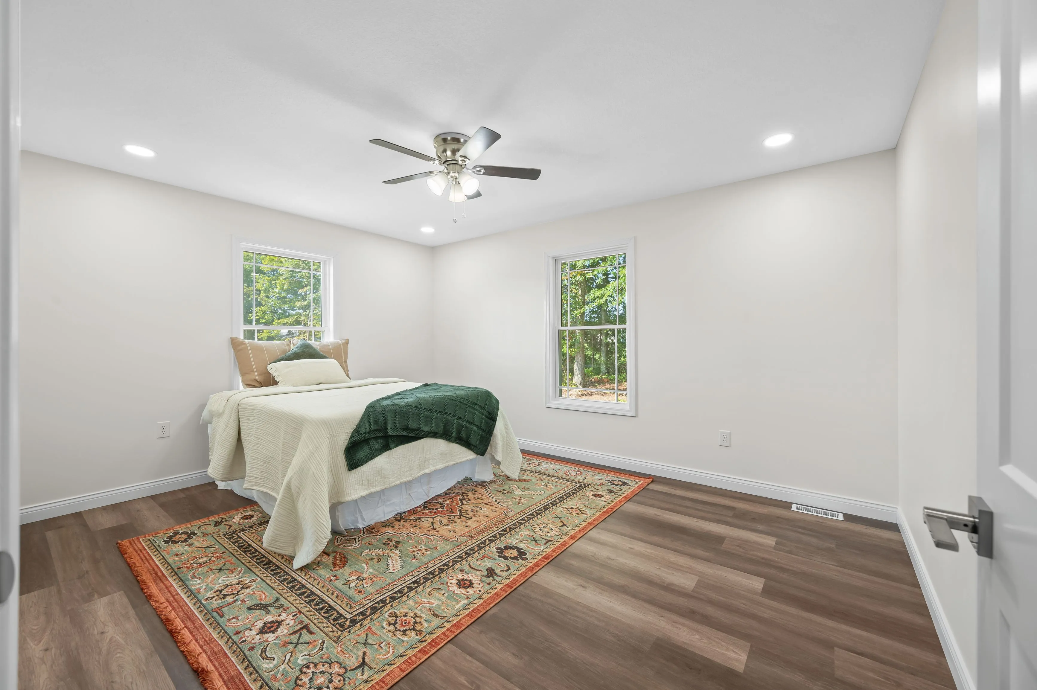 Bright minimalist bedroom with a bed, ceiling fan, recessed lighting, two windows, and a traditional area rug on a hardwood floor.