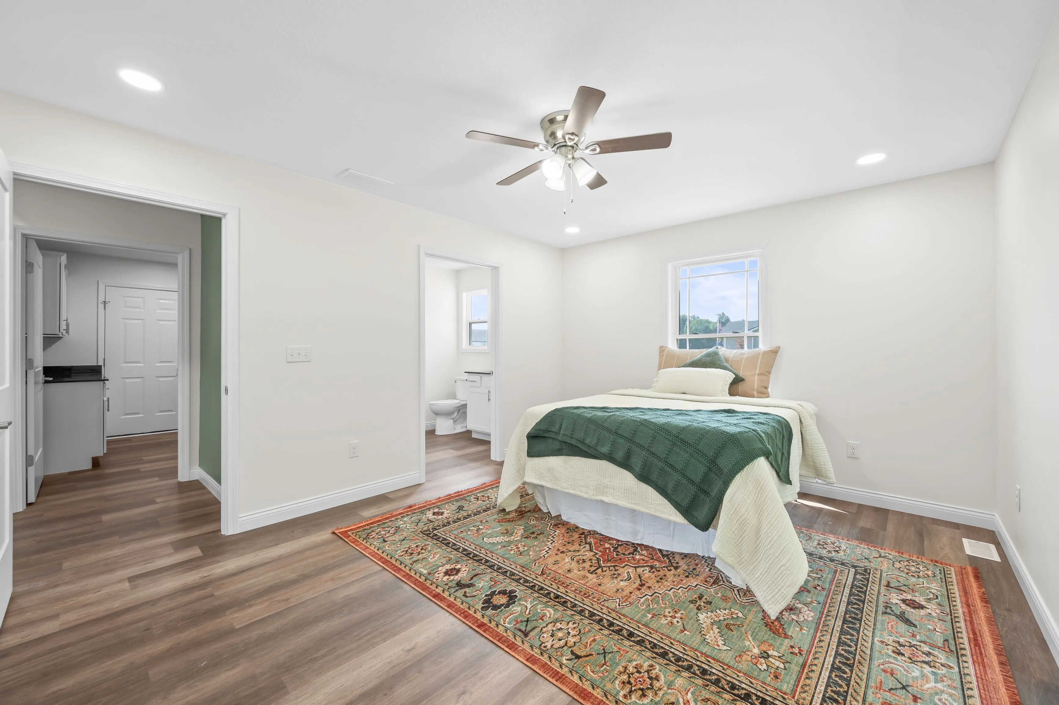 Spacious bedroom with large bed, green and white bedding, ornate area rug, ceiling fan, and ensuite restroom.