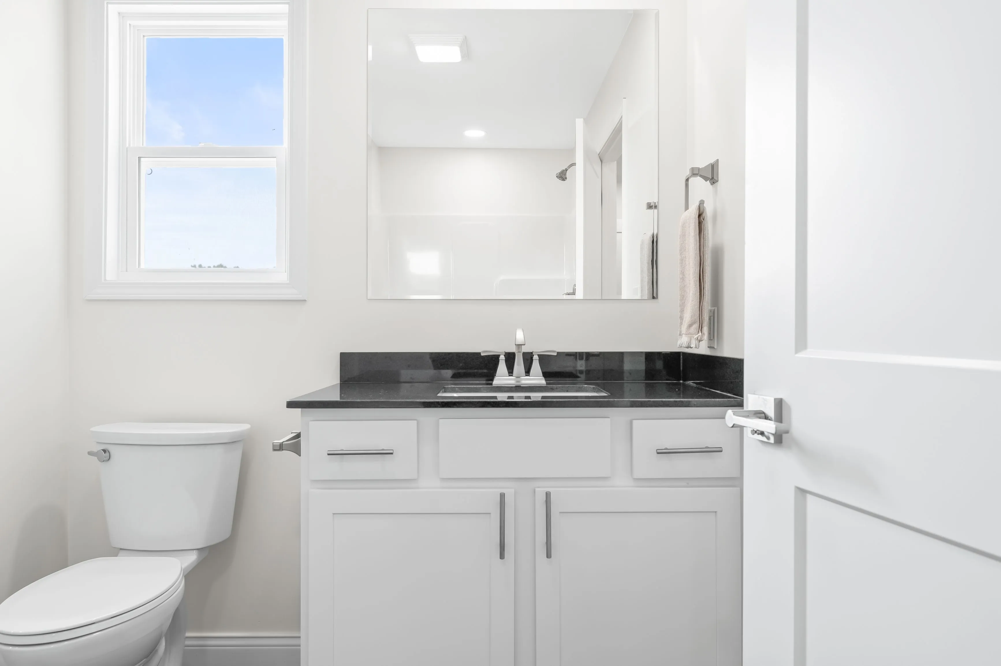 Modern bathroom interior with white walls, featuring a vanity with a black countertop, sink, mirror, and a toilet next to a window.