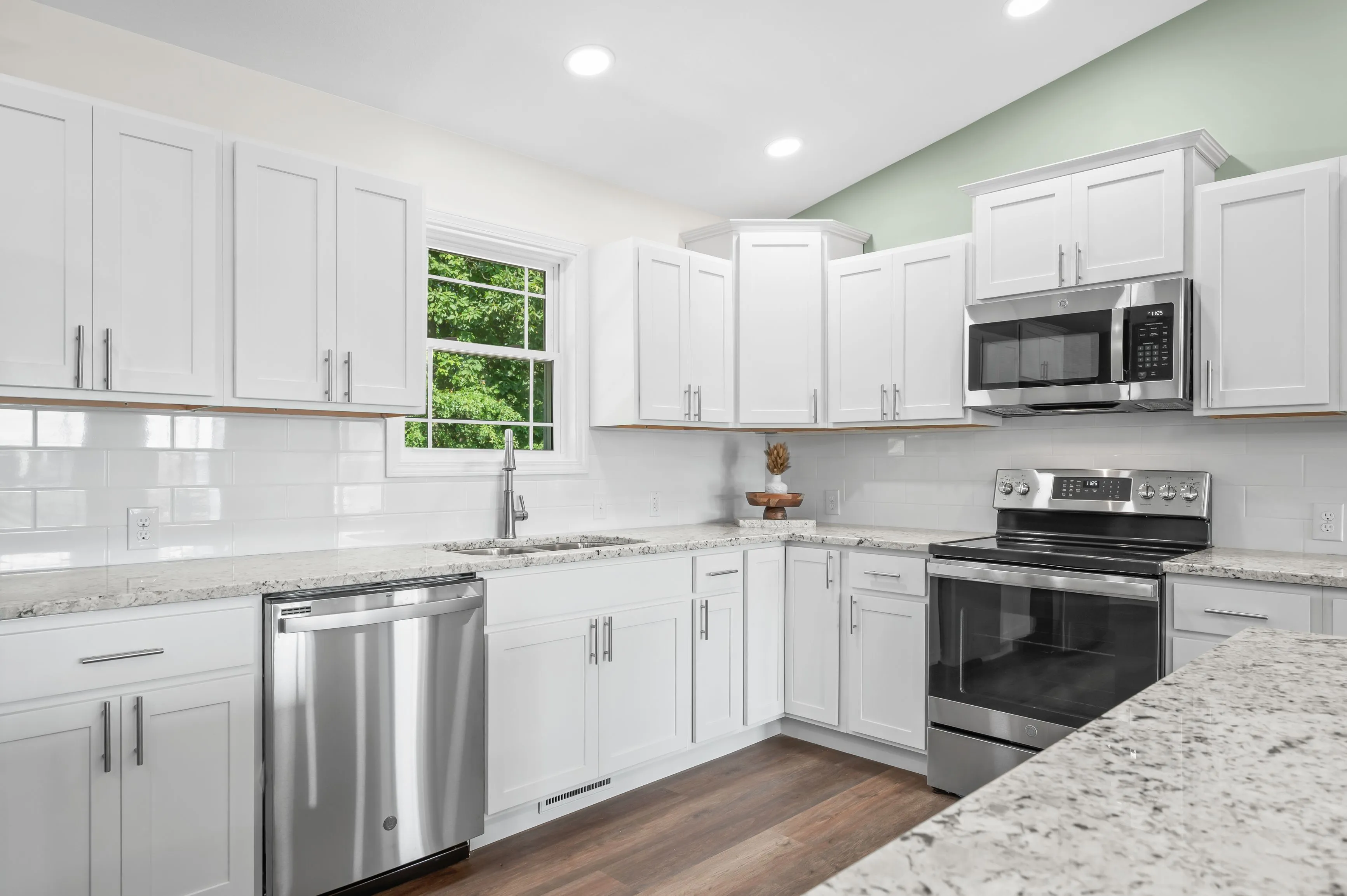 A modern kitchen interior with white cabinetry, stainless steel appliances, granite countertops, subway tile backsplash, and hardwood floors, with a view of greenery through a window.