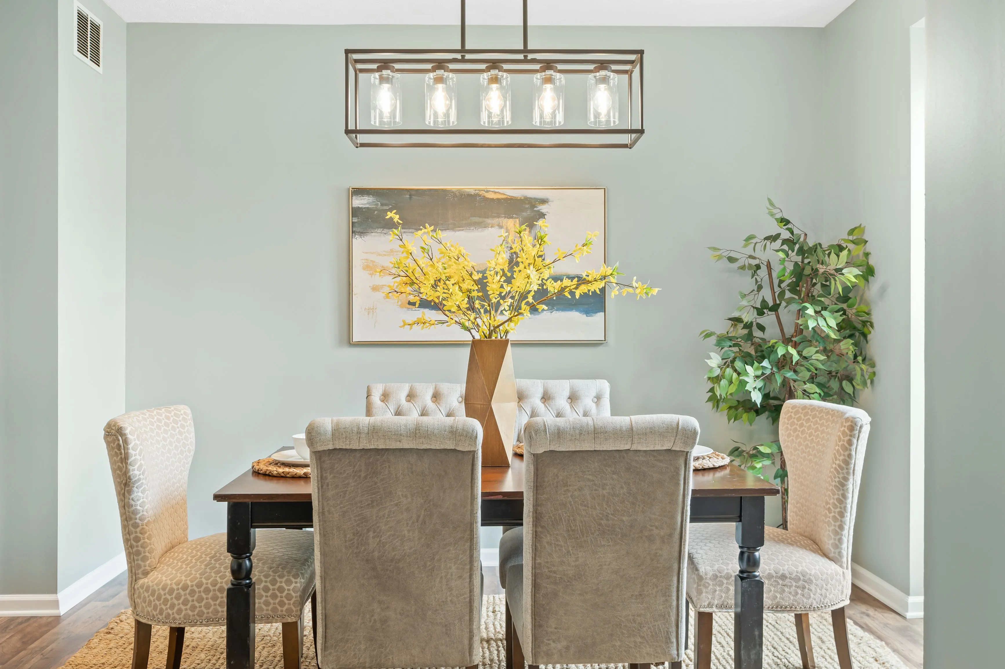Elegant dining room with a modern chandelier, a wooden table with mixed chair styles, and a painting of a tree above a console table.