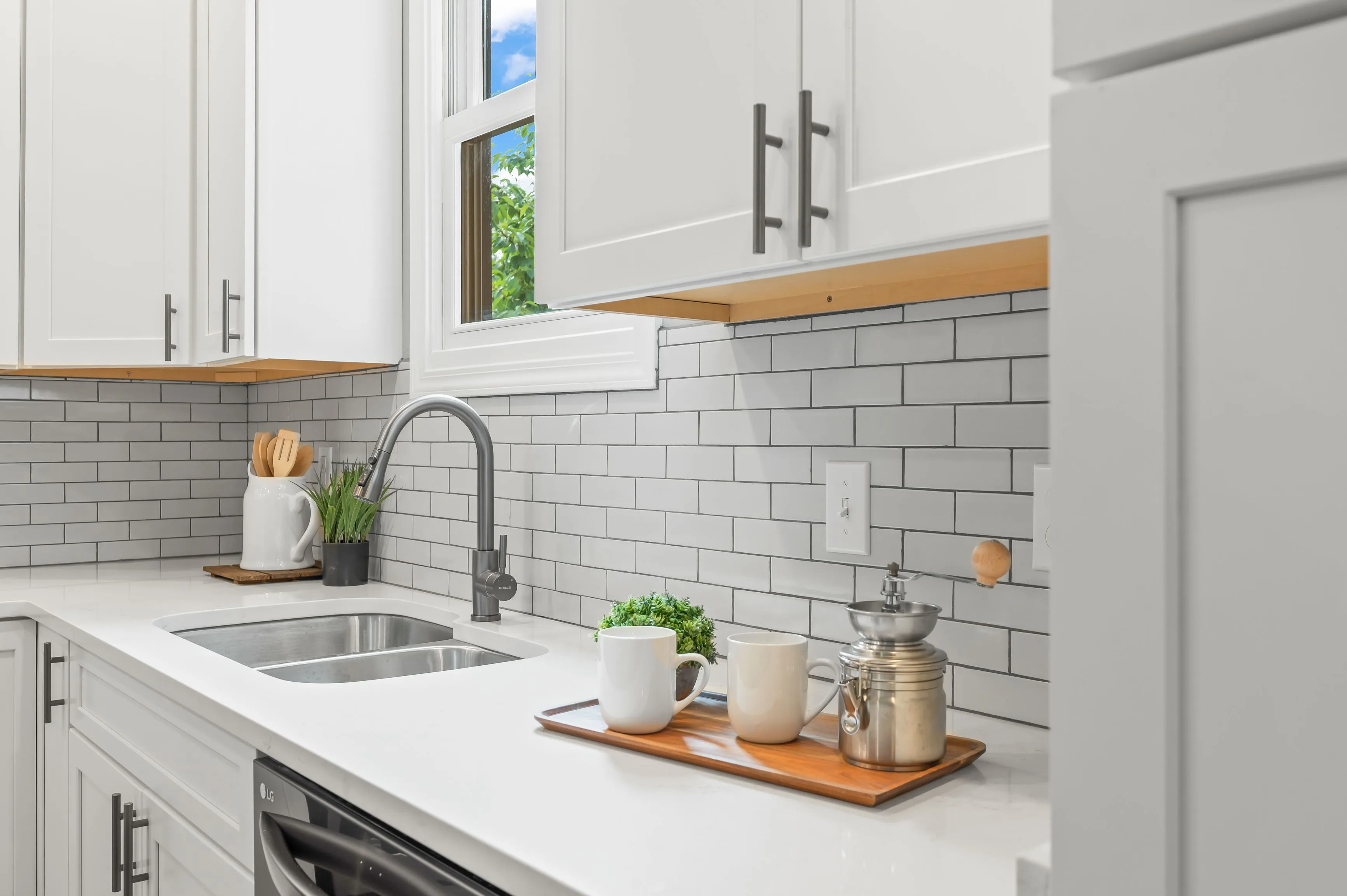 Bright modern kitchen with white cabinets, subway tile backsplash, and stainless steel sink under a window with greenery outside.