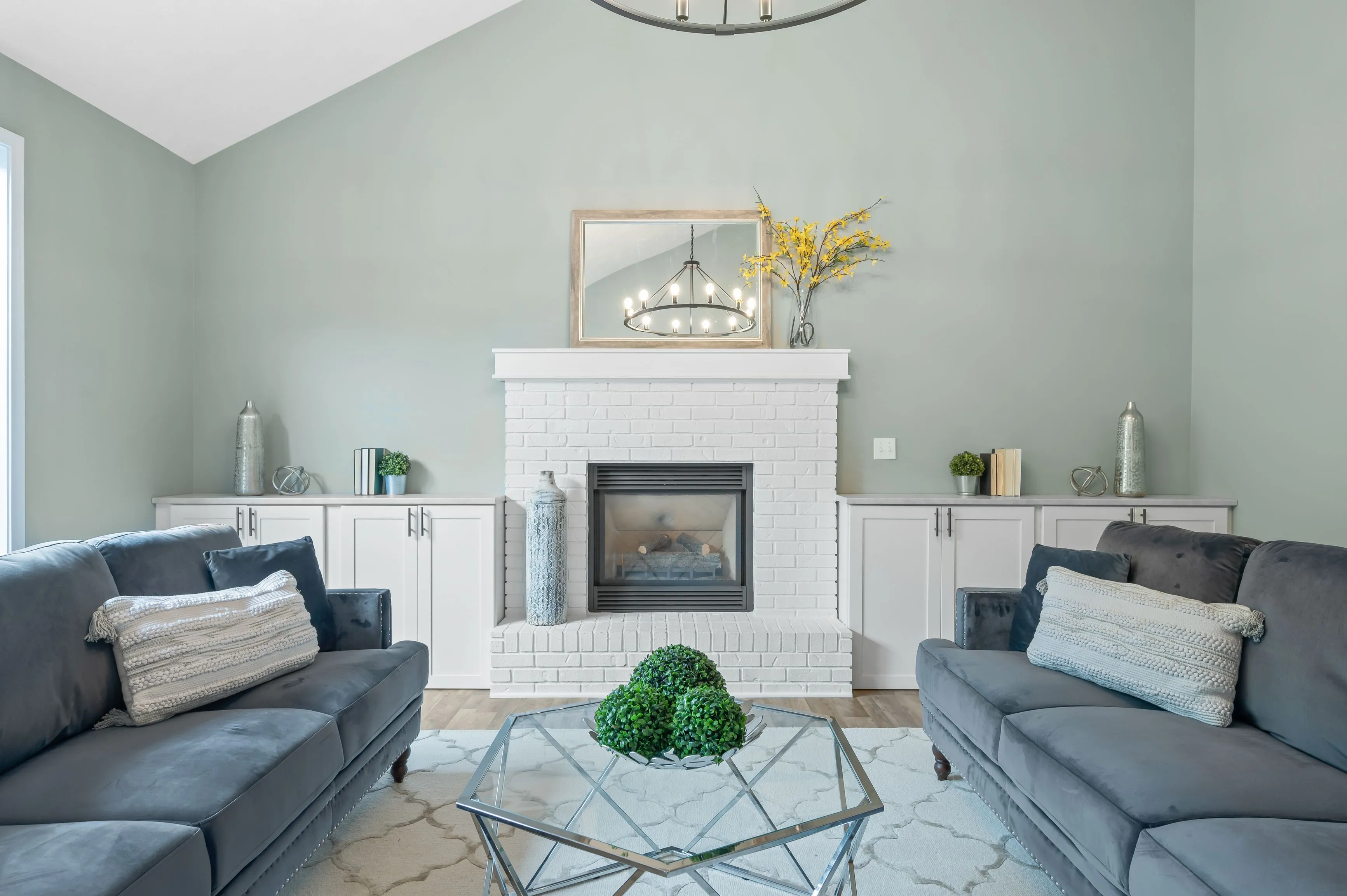 Elegant living room with light green walls, a white brick fireplace, two dark grey sofas, and a geometric glass coffee table with a decorative greenery centerpiece.