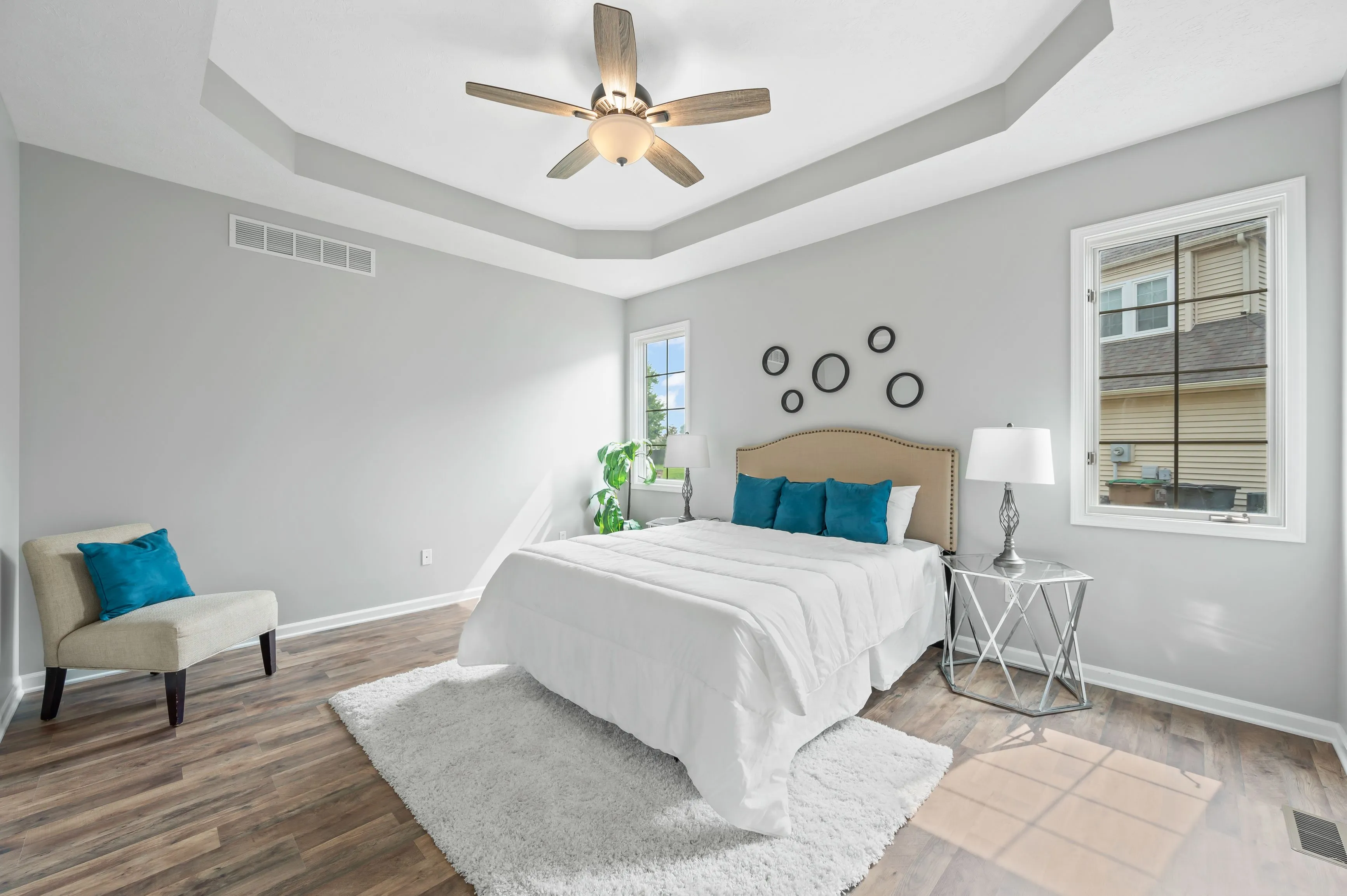 Bright modern bedroom with a large bed, ceiling fan, white walls with decorative mirrors, window with blinds, and a sitting area with a chair and side table.