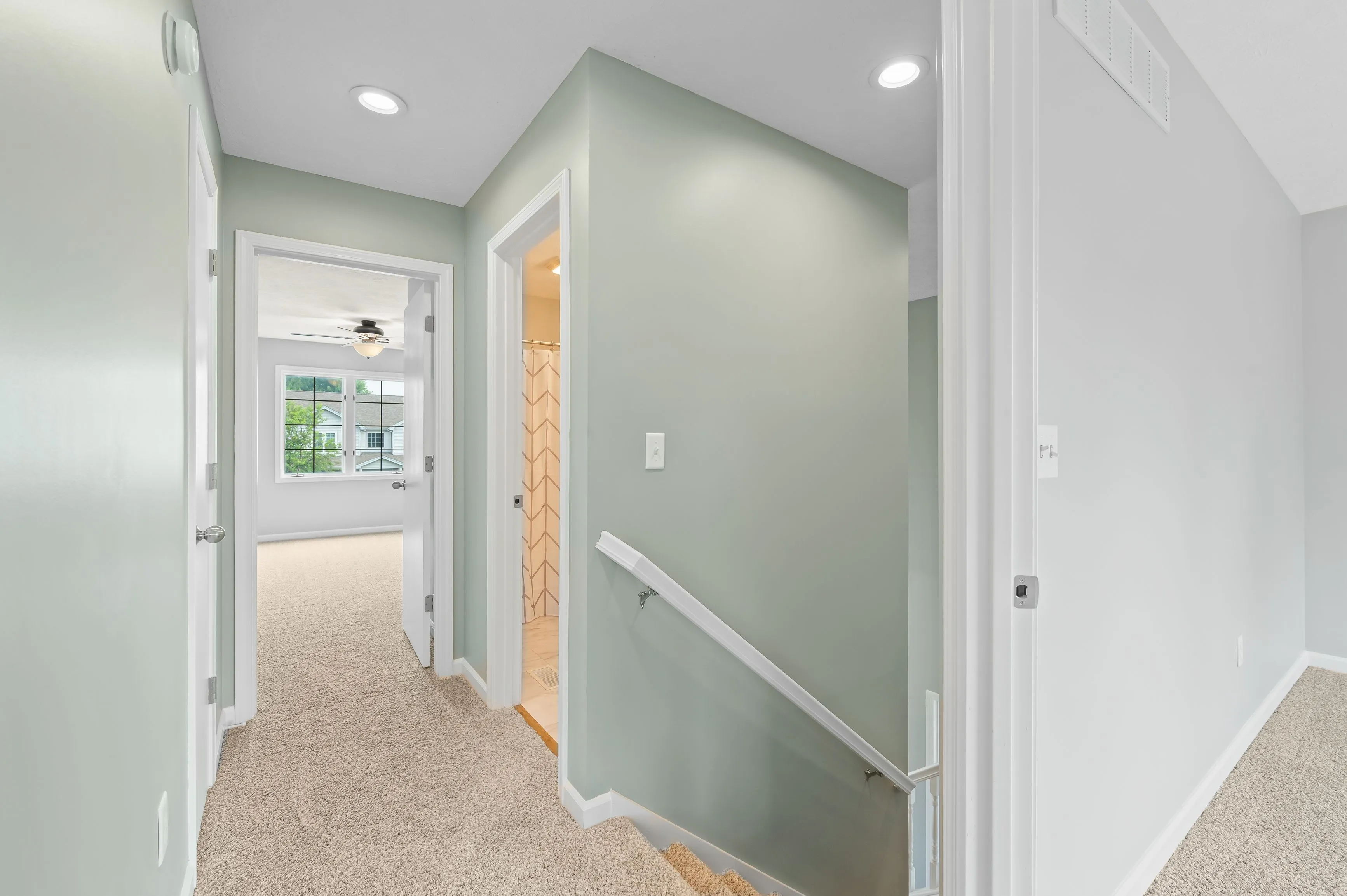 Bright hallway in a modern home with beige carpeting and white walls, featuring a railing and multiple doors leading to different rooms.