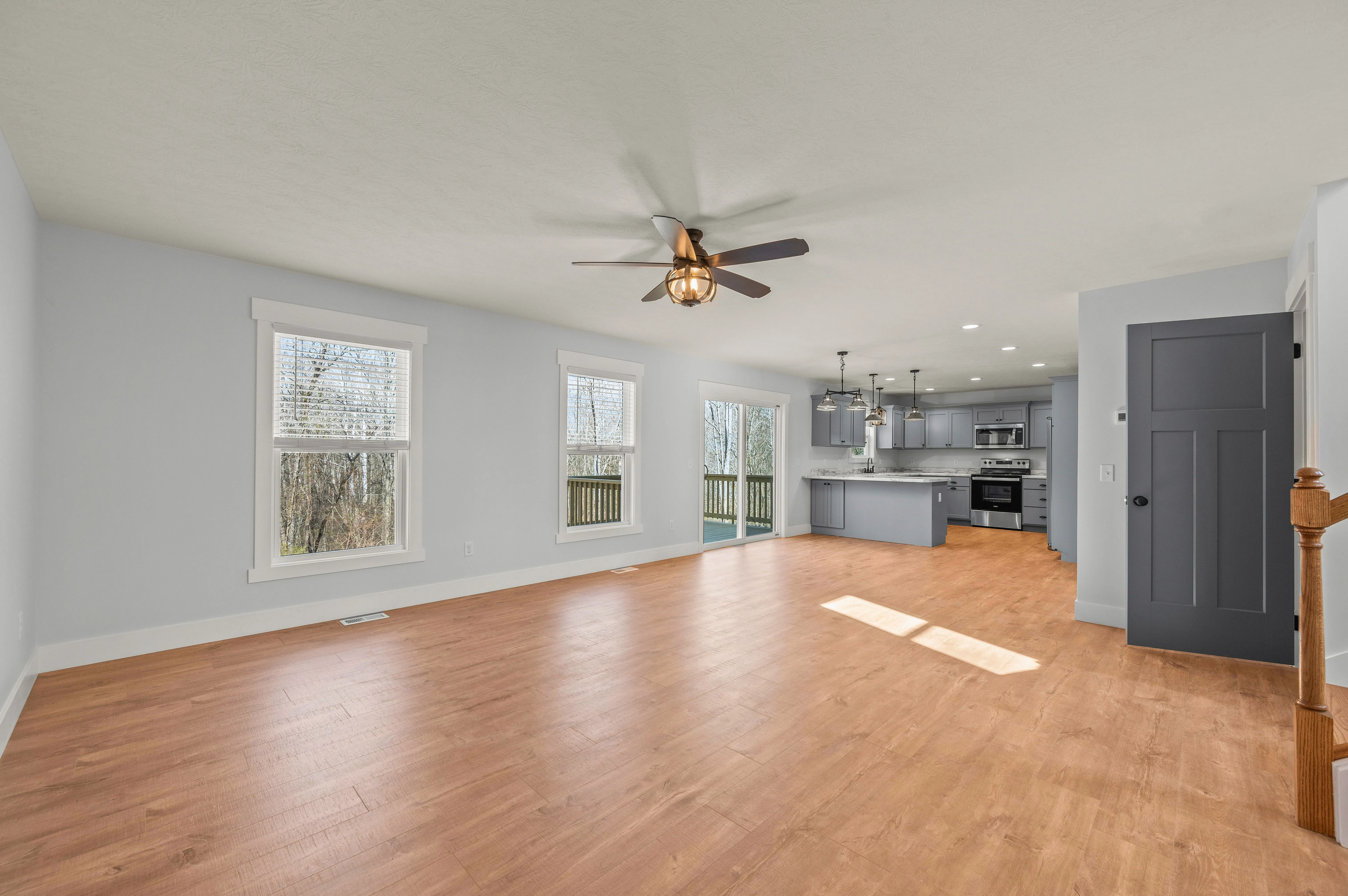 Spacious empty room with hardwood floors, pale blue walls, ceiling fan, and an open kitchen with modern appliances in the background.