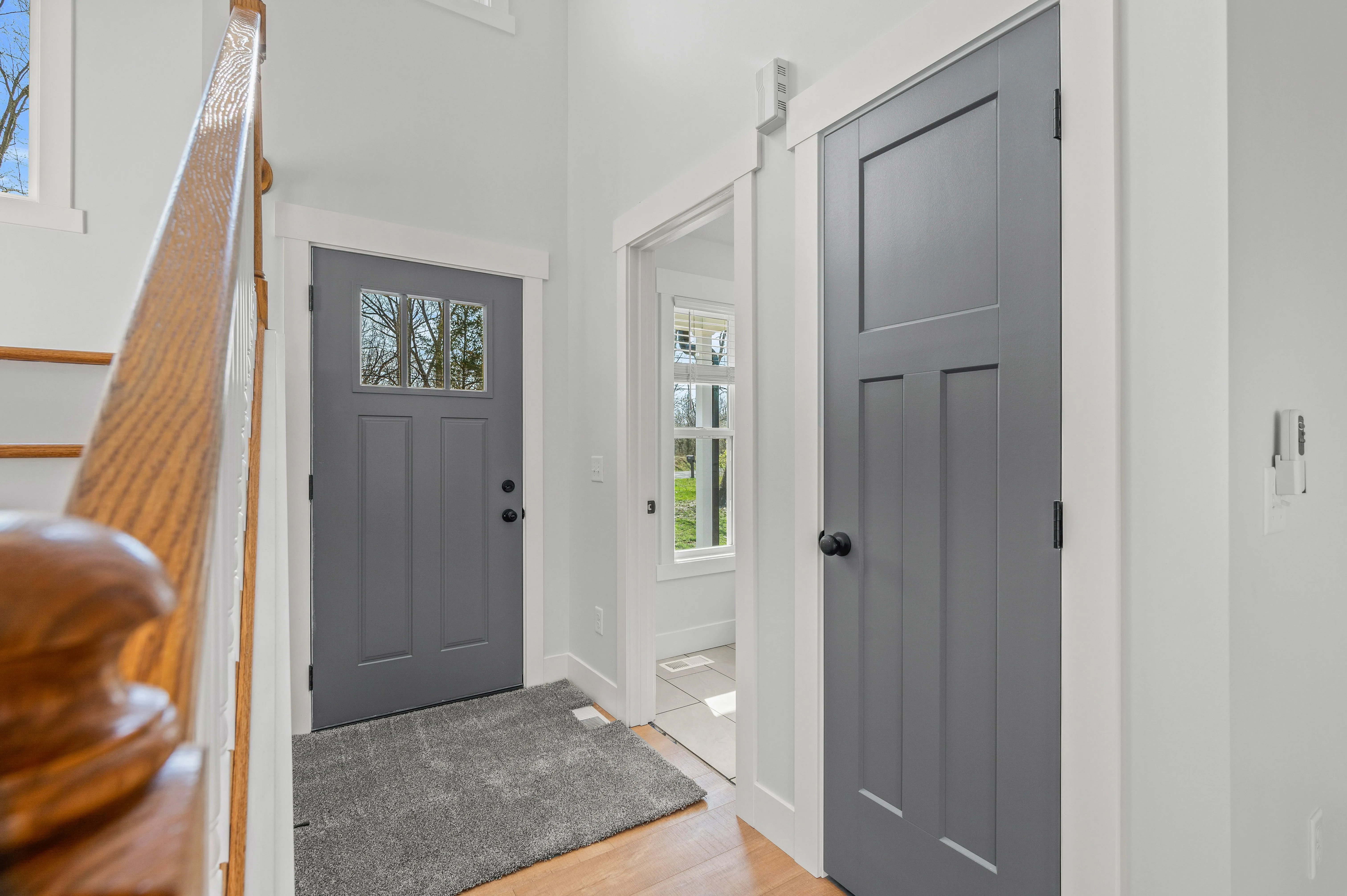 Bright interior hallway with wooden stair railing on the left, two gray doors, and a small window showing outdoor greenery.