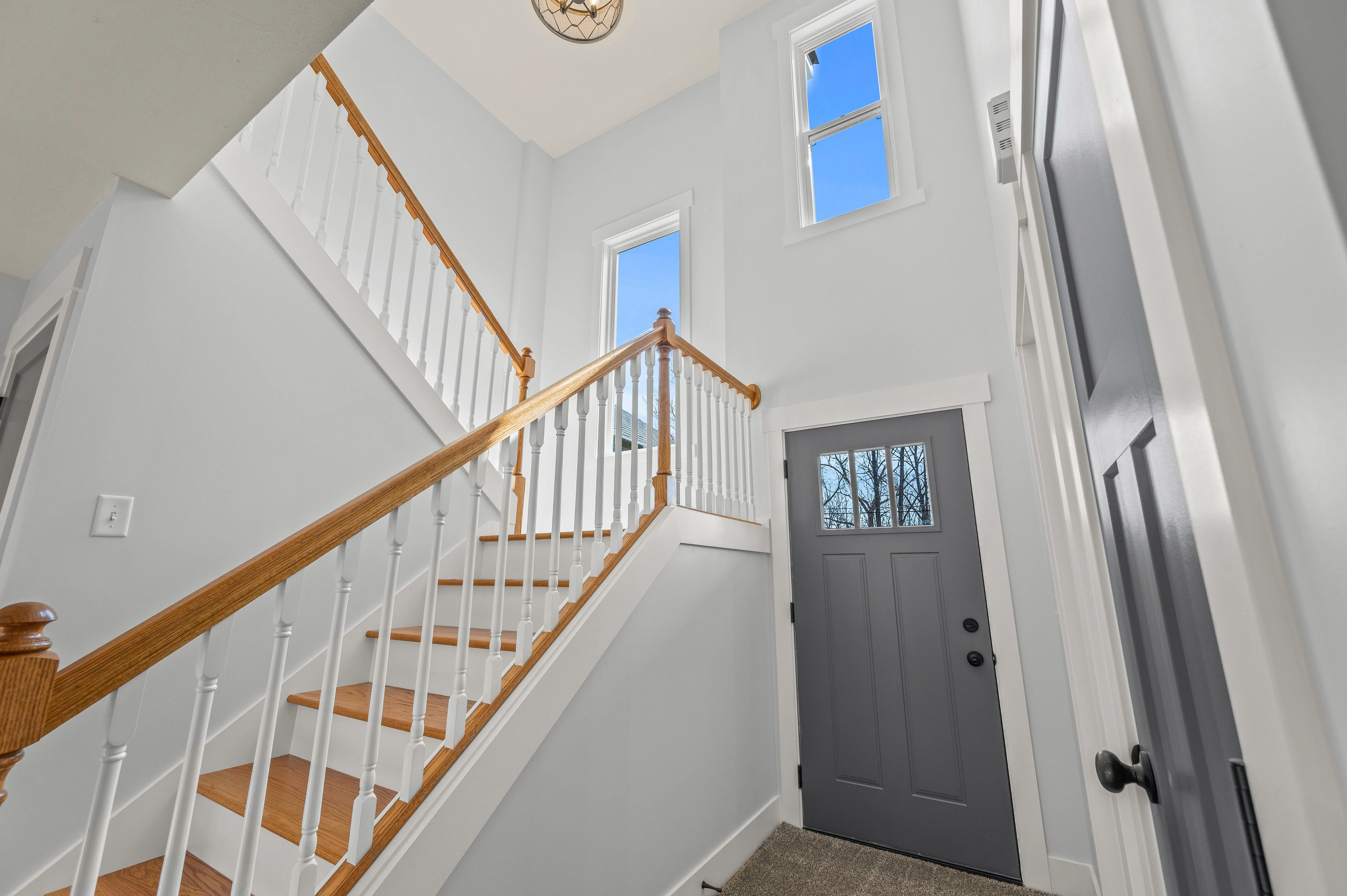 Bright entryway with staircase, wooden handrail, and white spindles, leading upstairs with a view of a gray front door and two windows.