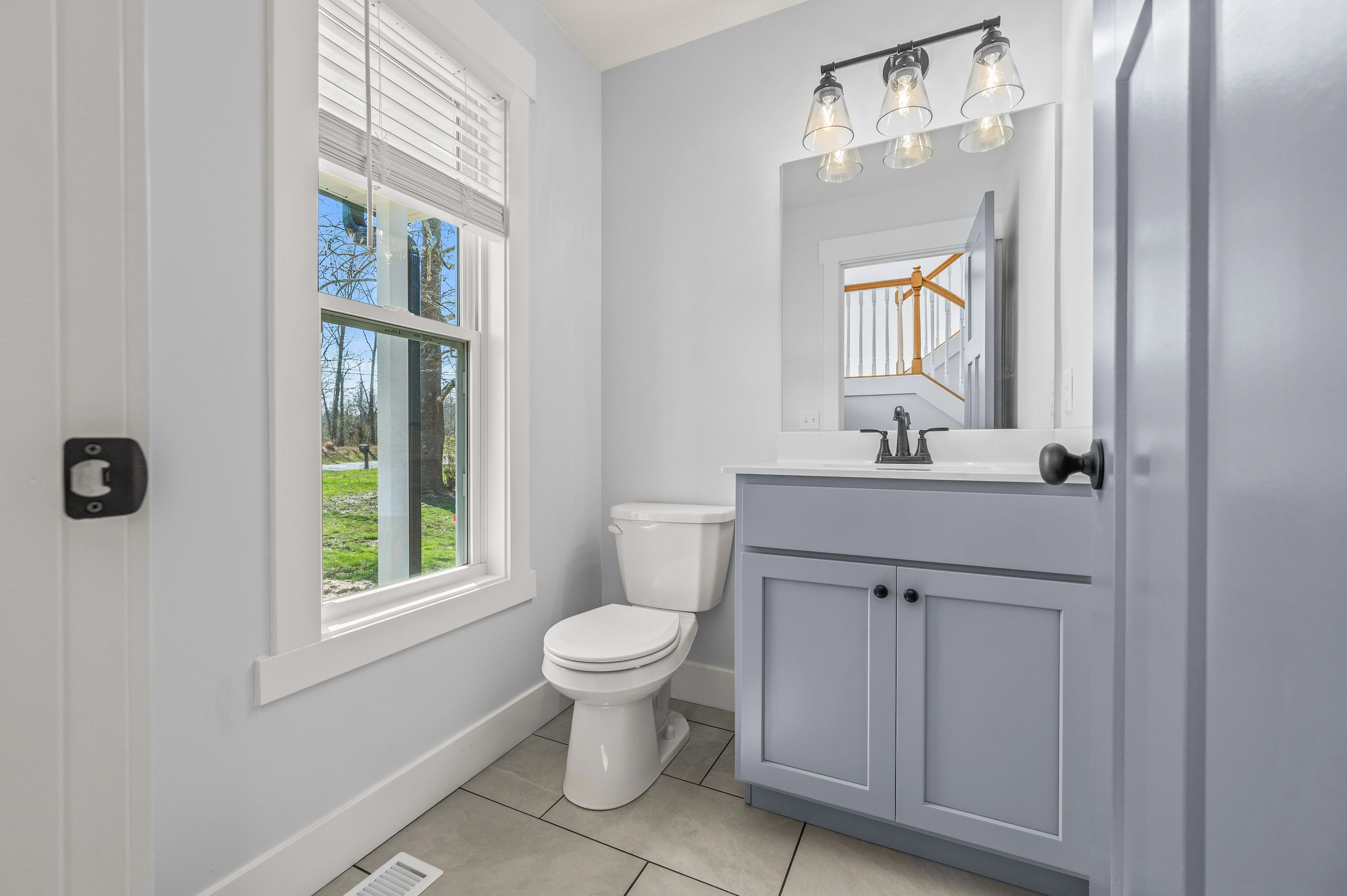 Modern bathroom interior with a window overlooking the yard, featuring a toilet and a blue vanity with sink and mirror.