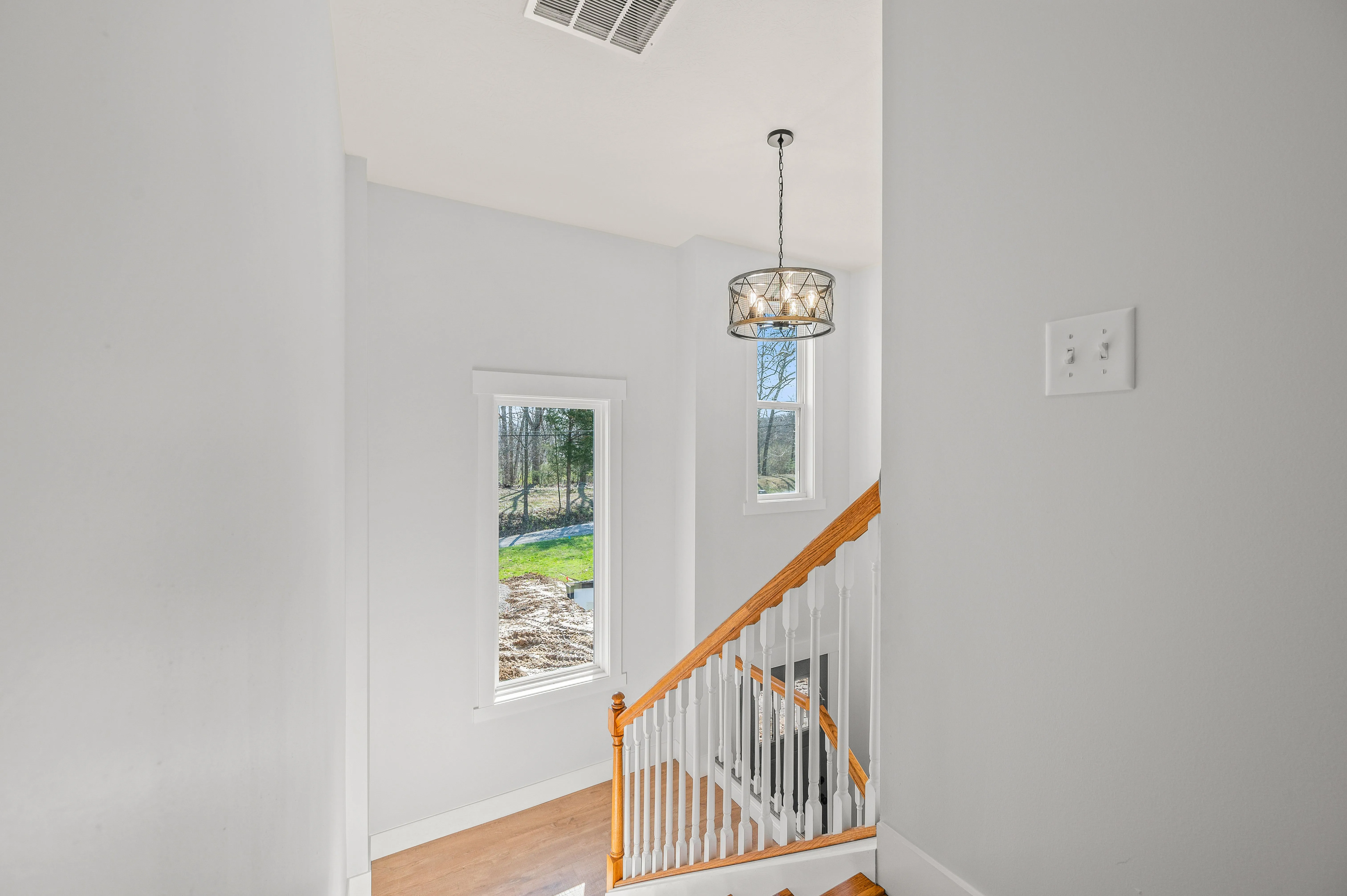 Brightly lit interior stairwell with wooden banister, geometric light fixture, and view of trees through window.