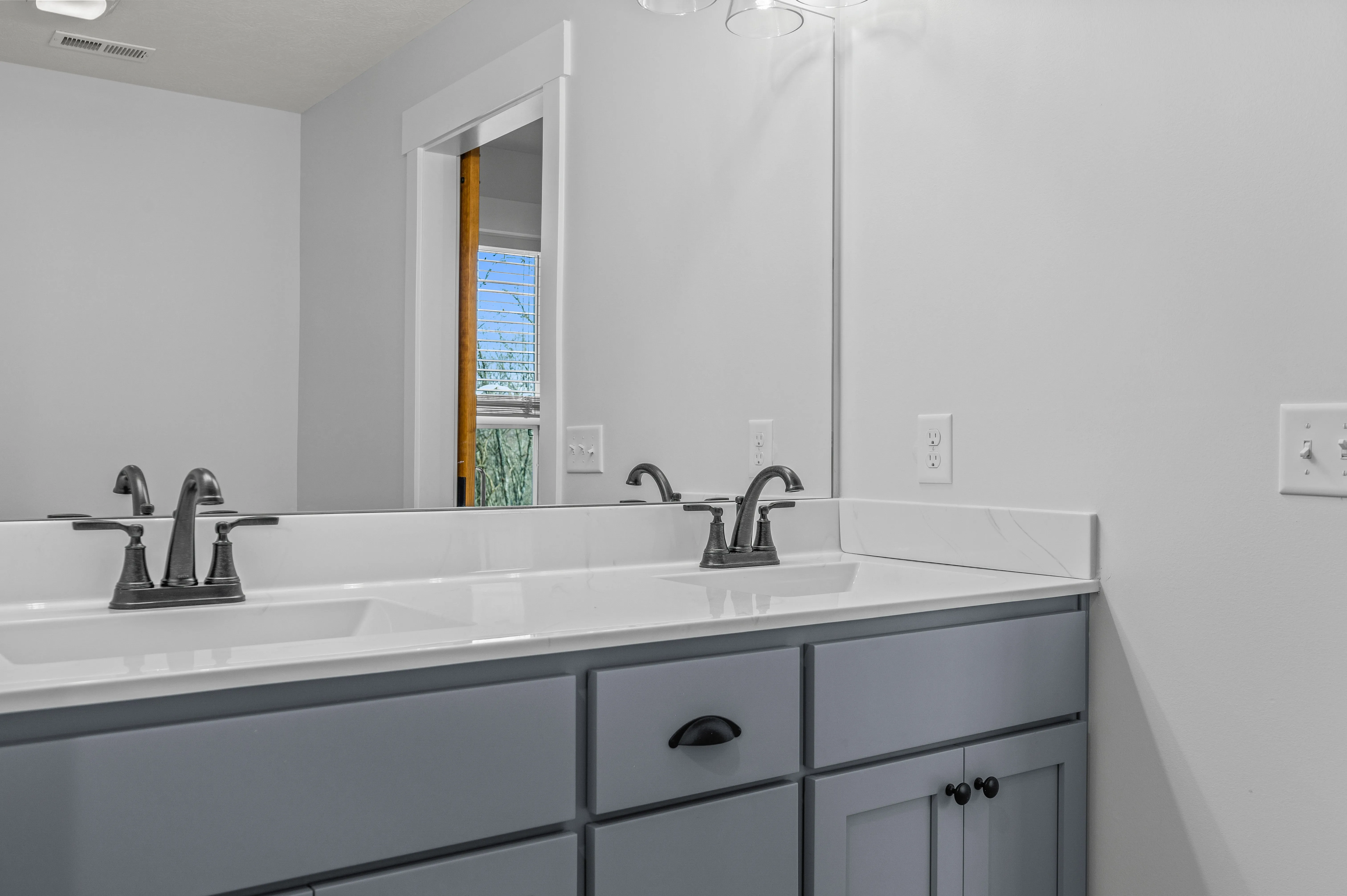 Modern bathroom interior with a double sink, gray vanity cabinet, and a mirror reflecting a window.