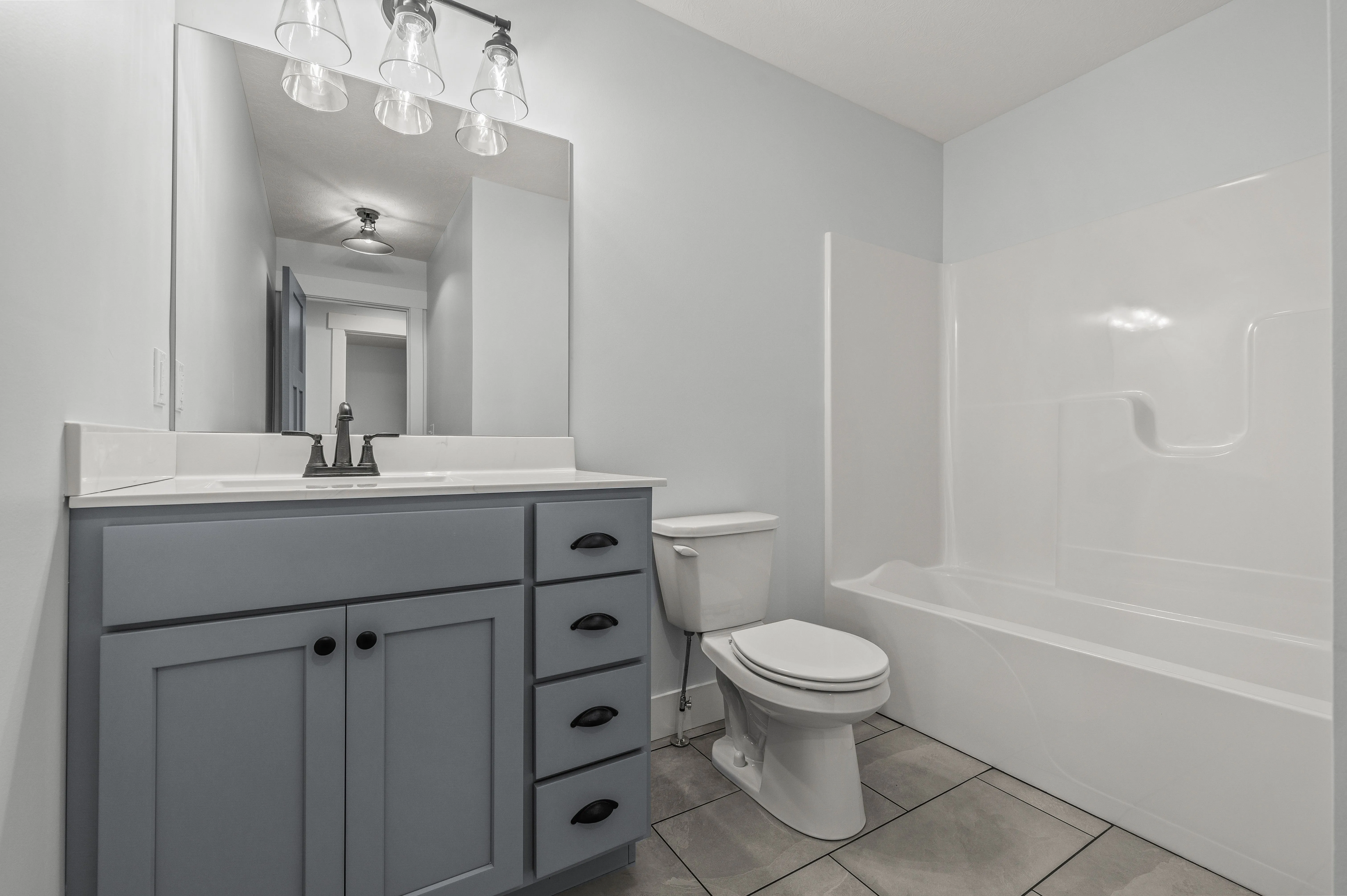 A modern bathroom interior with a gray vanity cabinet, a white countertop with a sink and faucet, a large mirror, a toilet, and a bathtub with a white surround.