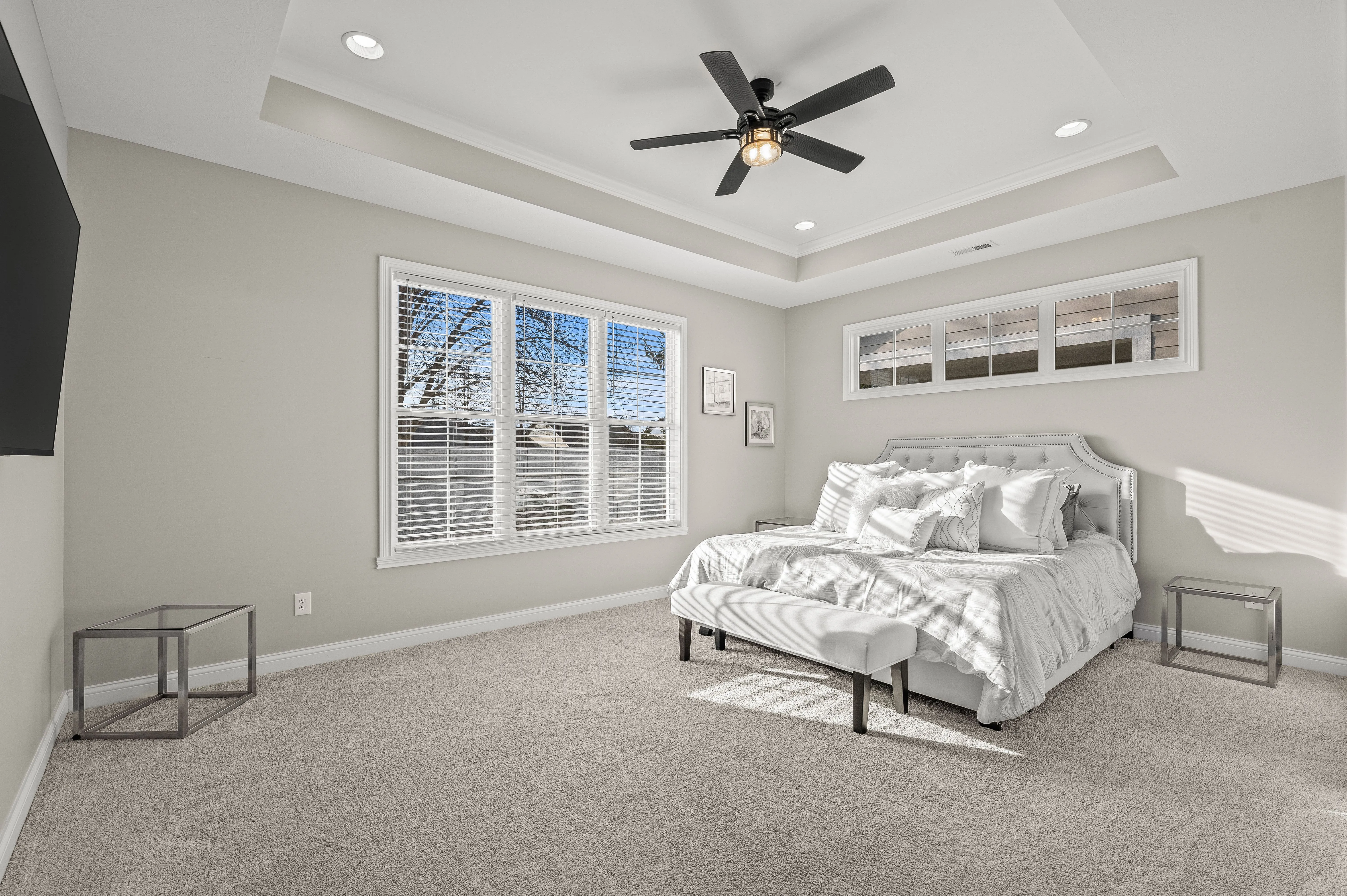 Modern bedroom with a white bed, carpeted floor, ceiling fan, and sunlight shining through windows.