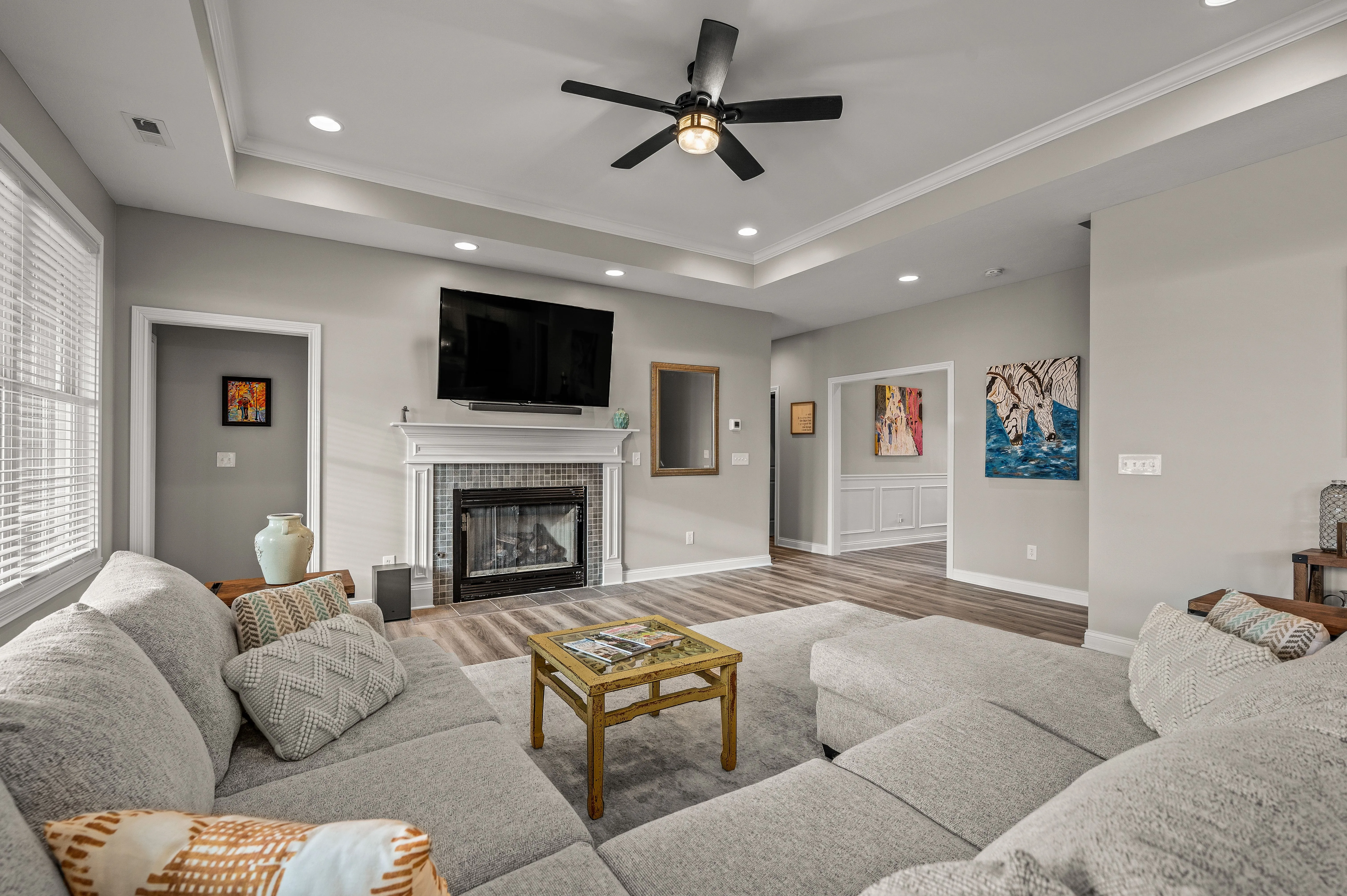 Spacious modern living room with gray sectional sofa, hardwood floors, white fireplace, ceiling fan, and wall-mounted television.